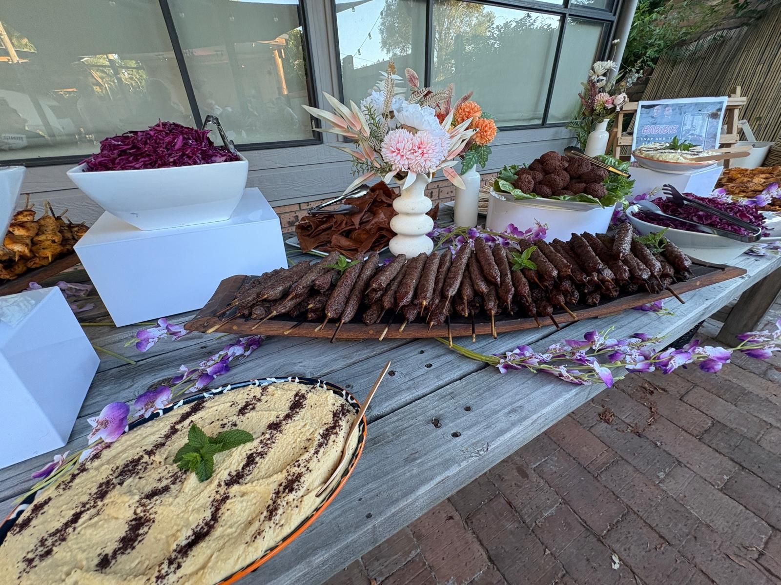 Buffet table with various dishes: hummus, kebabs, salad, and flowers. Outdoor setting. — The Habibiz Catering In Wingham, NSW