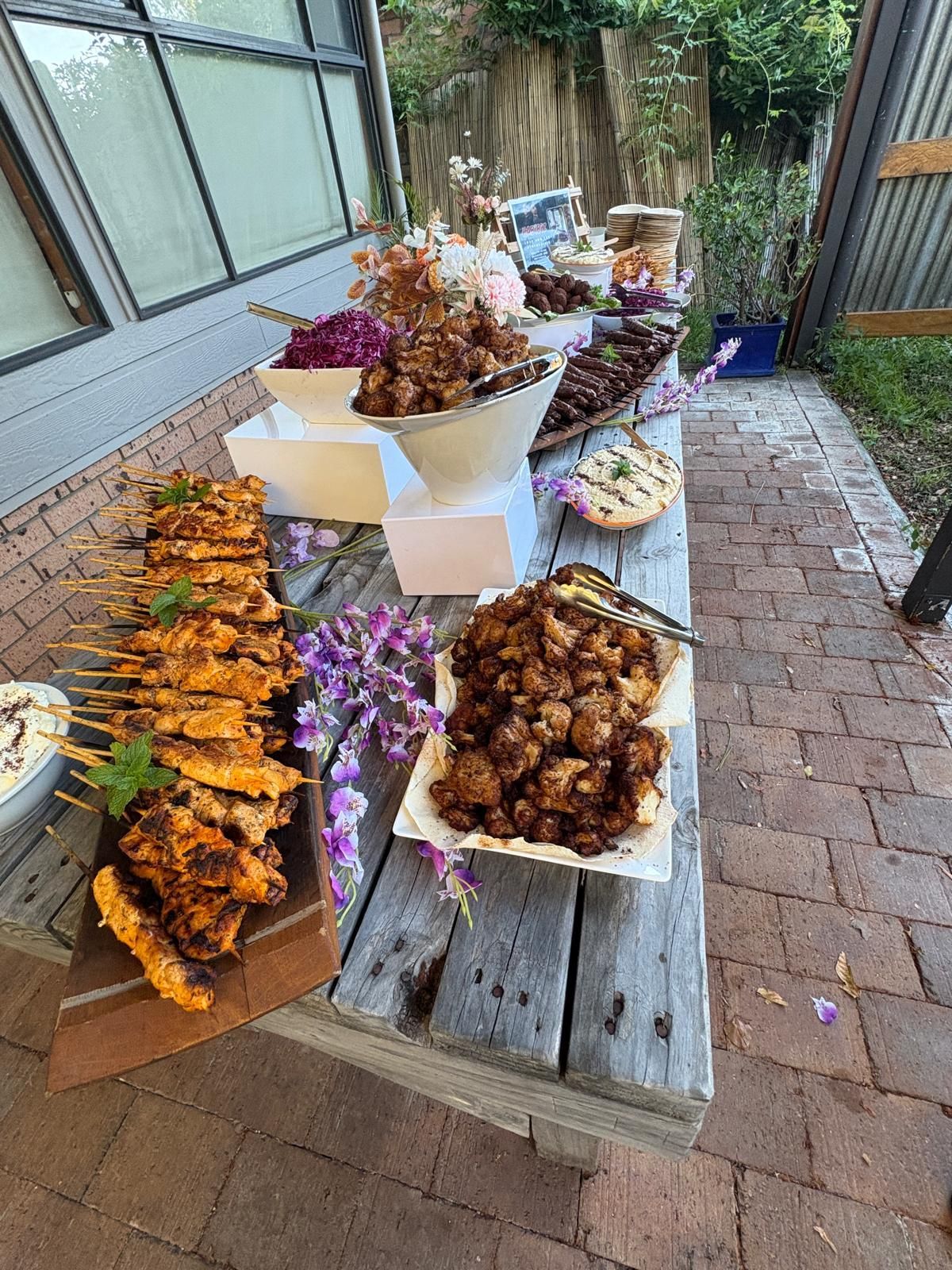 Food buffet on weathered wooden table, skewers, fried items, and decorative flowers, outdoor setting. — The Habibiz Catering In Wingham, NSW