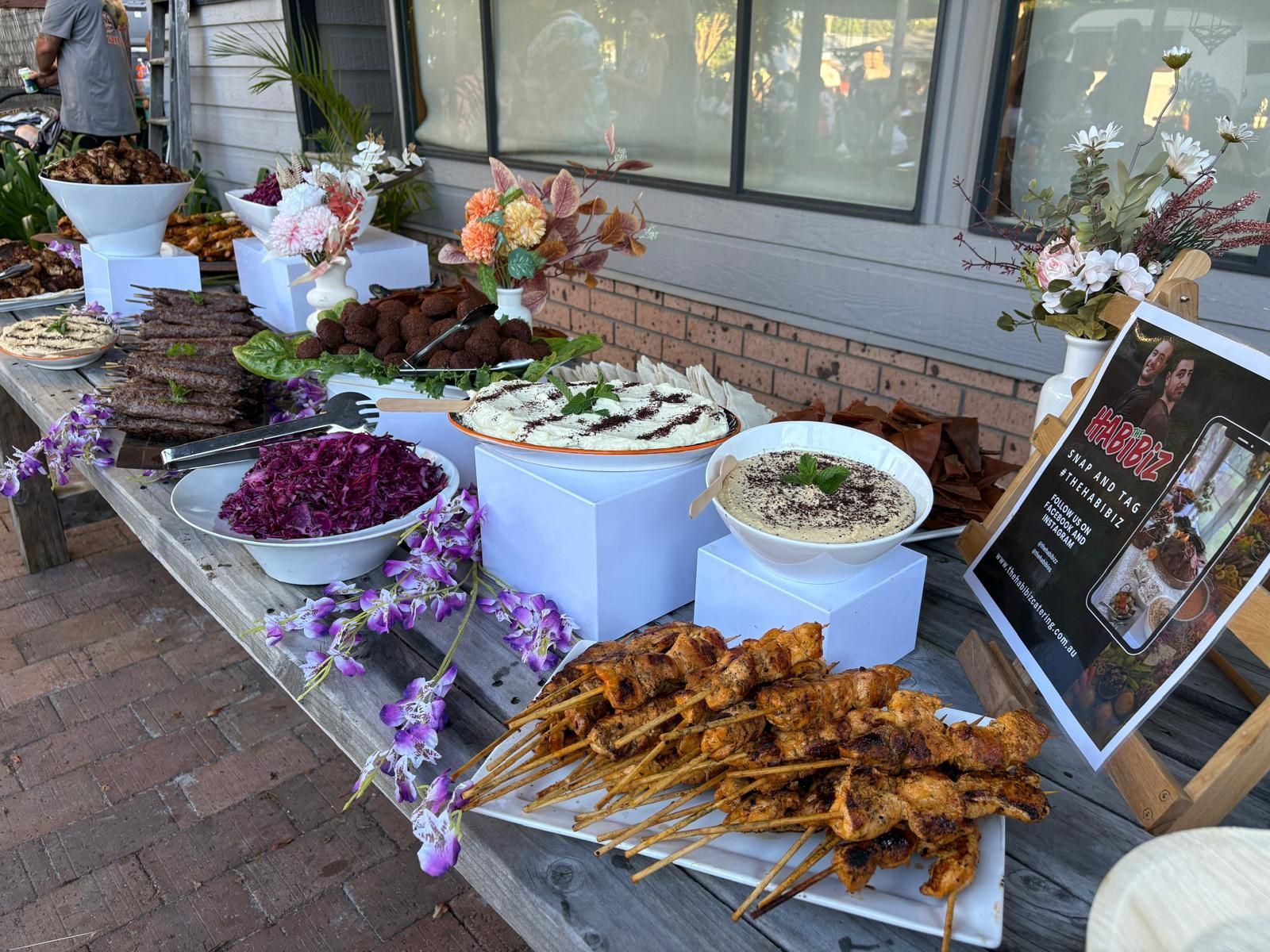 Food buffet with skewers, dips, and falafel, arranged on a long wooden table. White floral arrangements and a sign are present. — The Habibiz Catering In Wingham, NSW