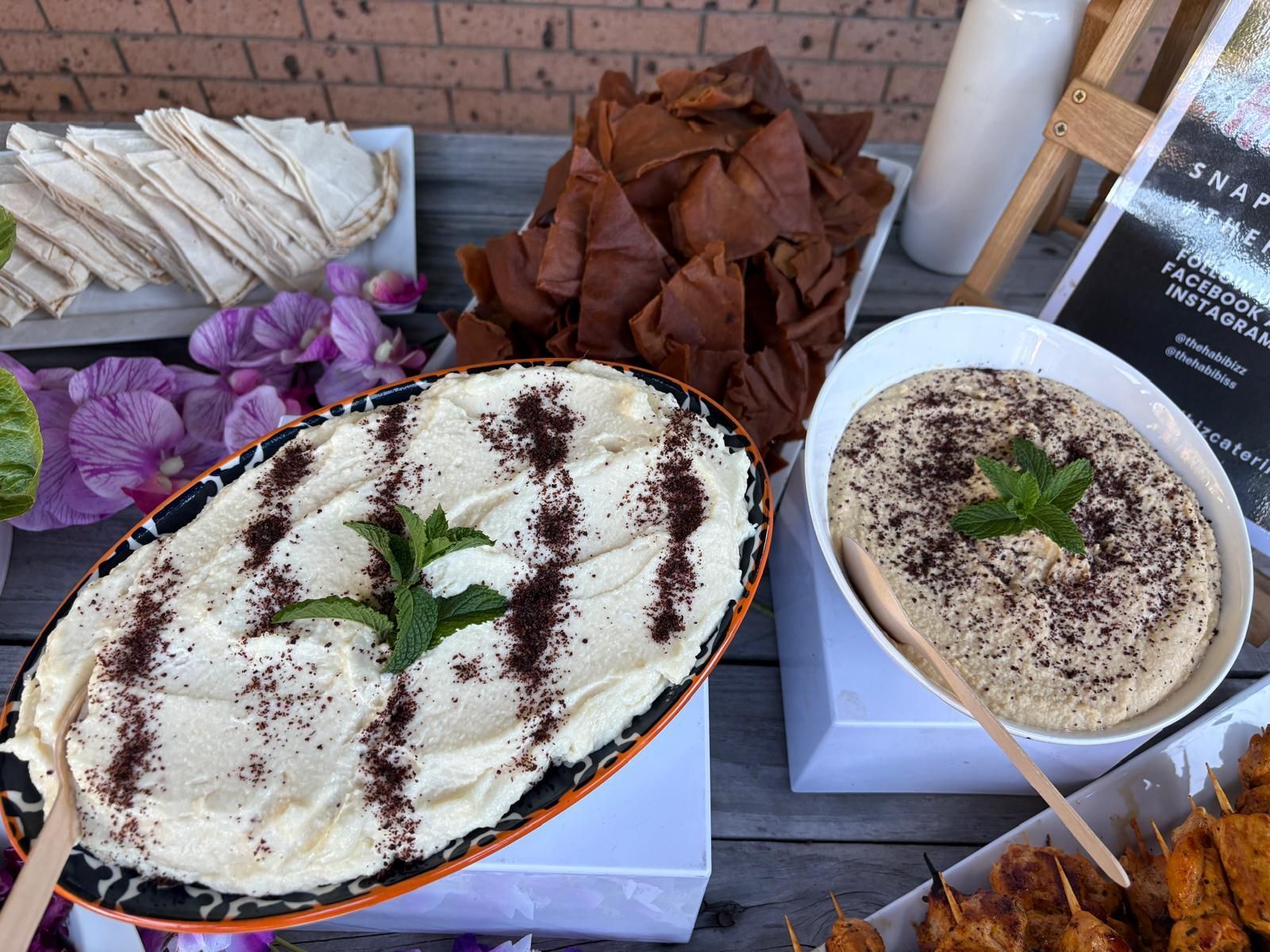 Hummus platter with pita bread, and toasted pita chips, with purple flowers and a sign on a table. — The Habibiz Catering In Wingham, NSW