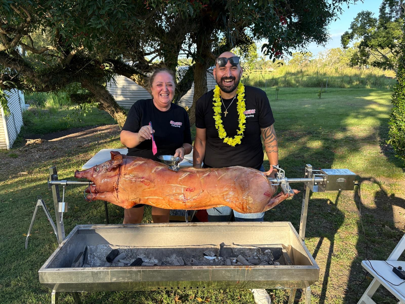 Two people stand behind a roasted pig on a spit over a charcoal grill outdoors.— The Habibiz Catering In Wingham, NSW