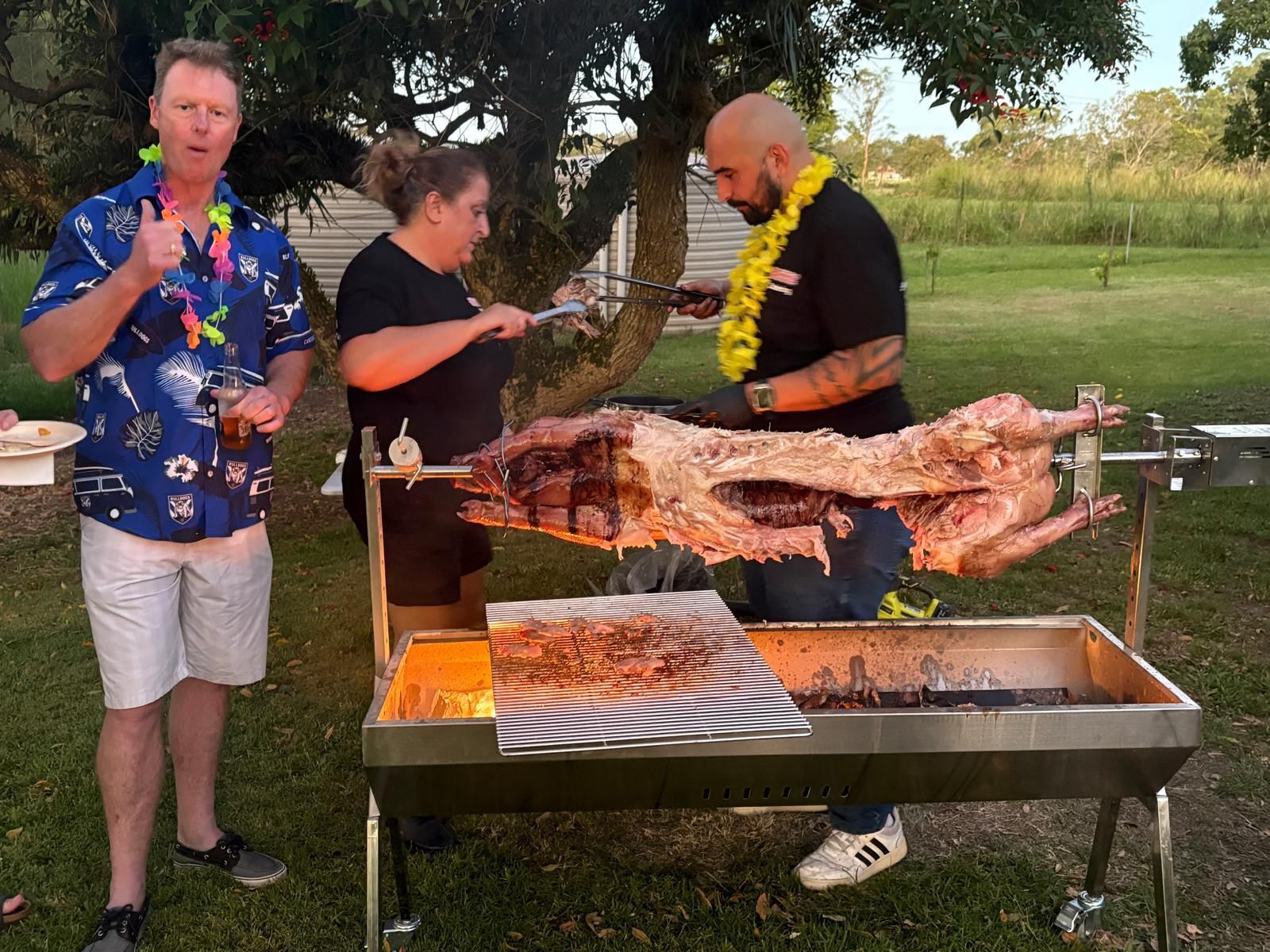Three people roasting a whole animal over a fire pit outdoors; one gives a thumbs up. — The Habibiz Catering In Wingham, NSW