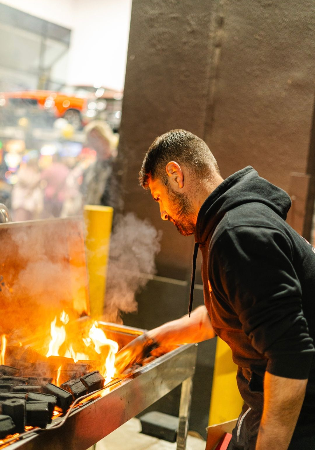 Man with beard tending to a fiery grill, indoors. He's wearing a black hoodie, smoke rising. - The Habibiz Catering in Wingham NSW