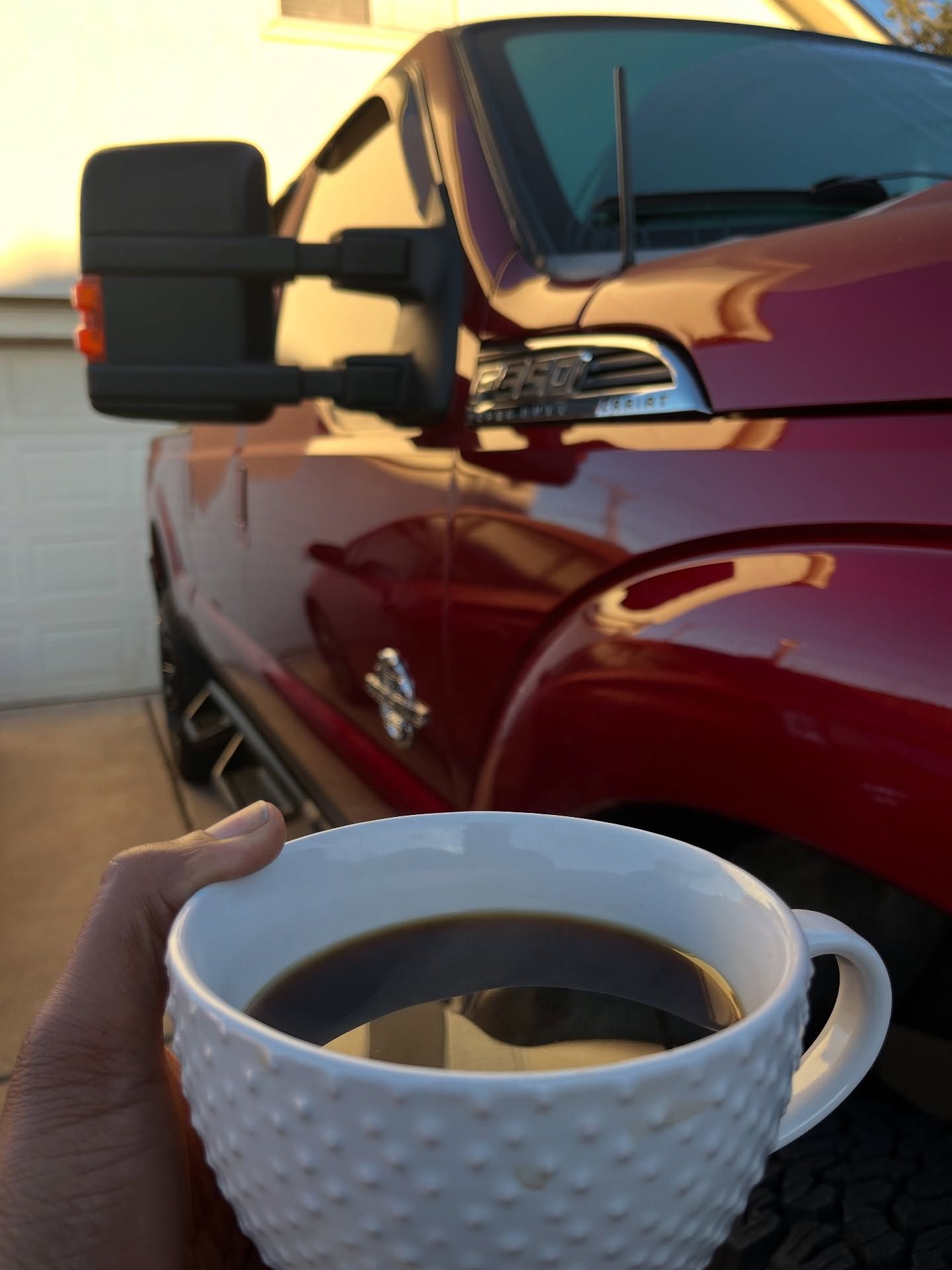 Hand holding a white textured mug with coffee in front of a red truck.