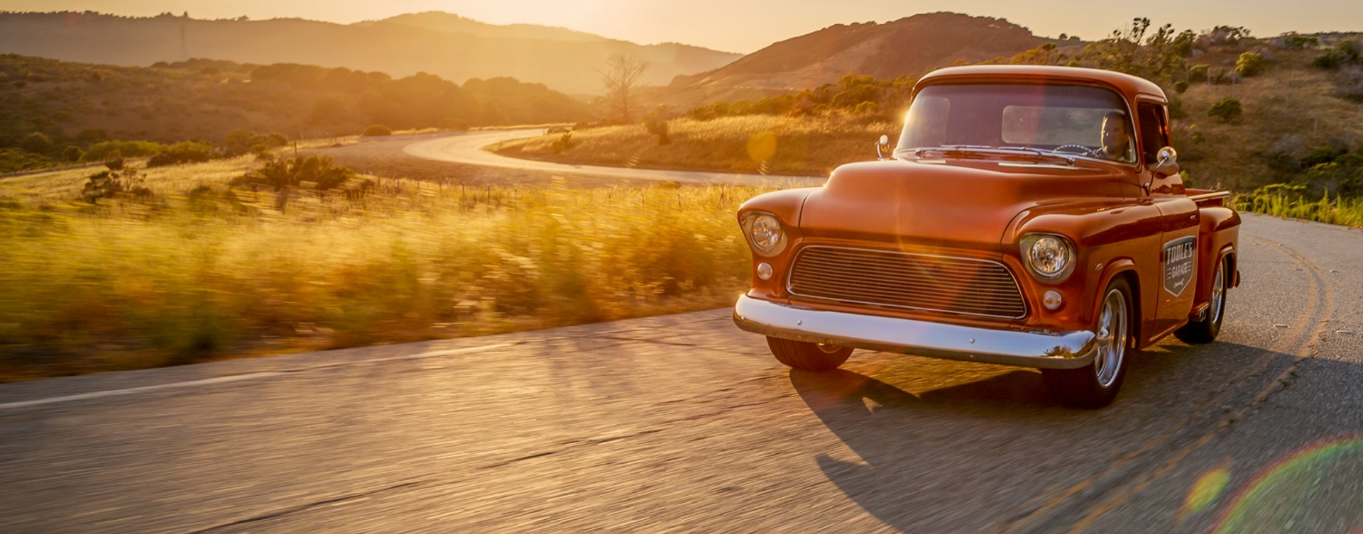 An old red truck is driving down a country road at sunset.