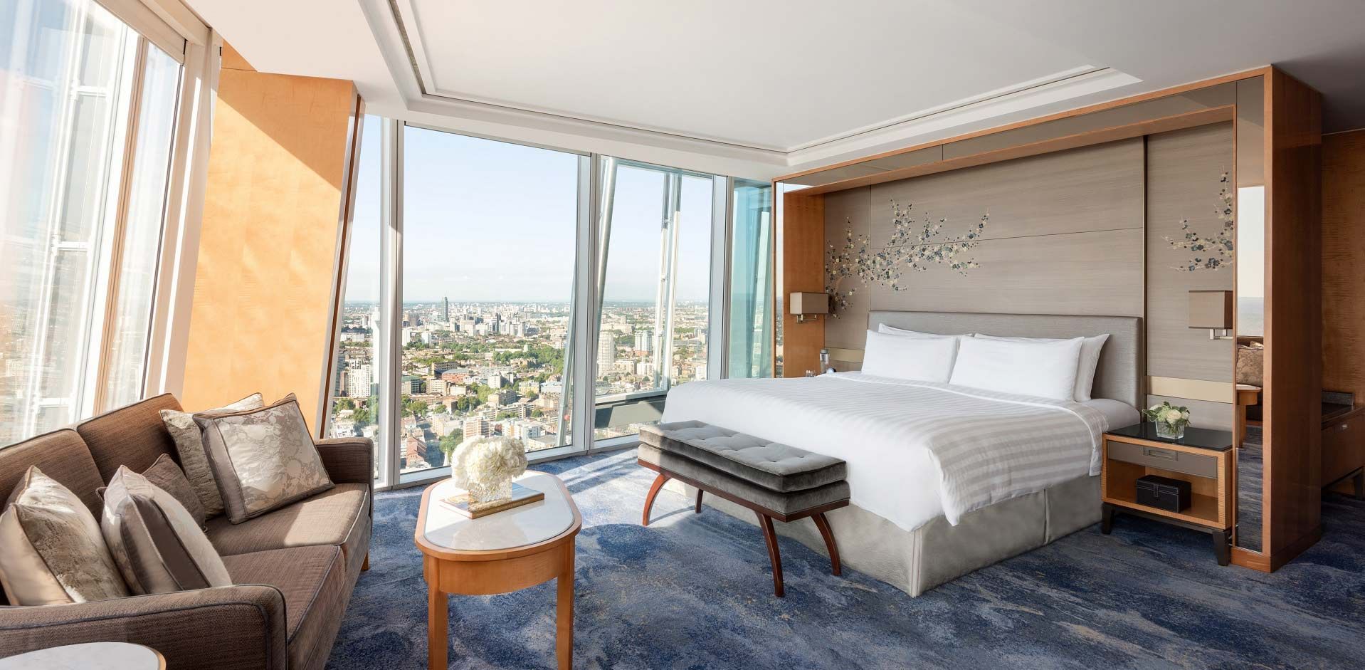 Um quarto de hotel moderno com uma cama grande, um sofá e janelas panorâmicas com vista para o horizonte da cidade.