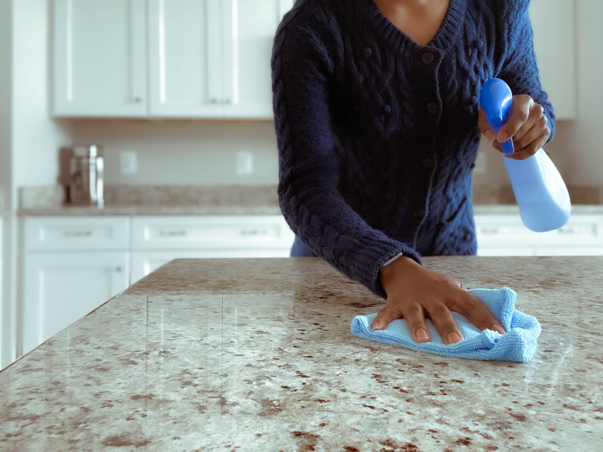 A woman is cleaning a granite counter top with a cloth and spray bottle.