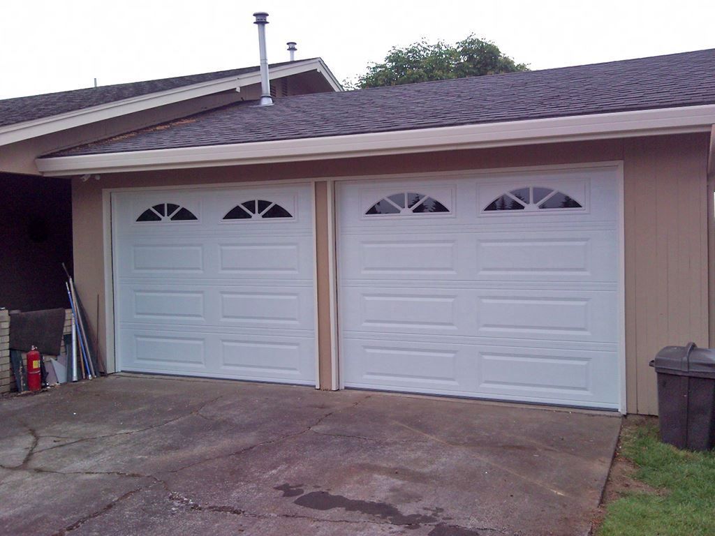 Two white garage doors with arched windows, tan building, asphalt driveway, and a trash can.