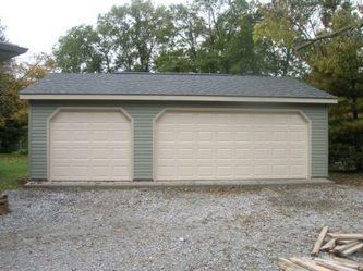 Two-car garage with light-colored doors and green siding. Gravel driveway, trees in the background.