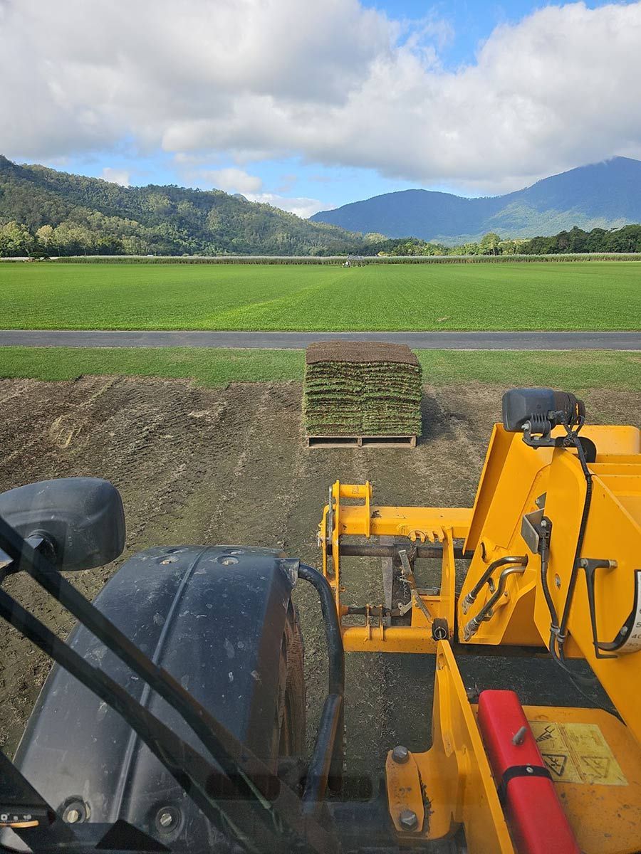A Yellow Tractor Is Driving Through A Grassy Field With Mountains In The Background — Tropical Lawns Pty Ltd In Little Mulgrave Qld