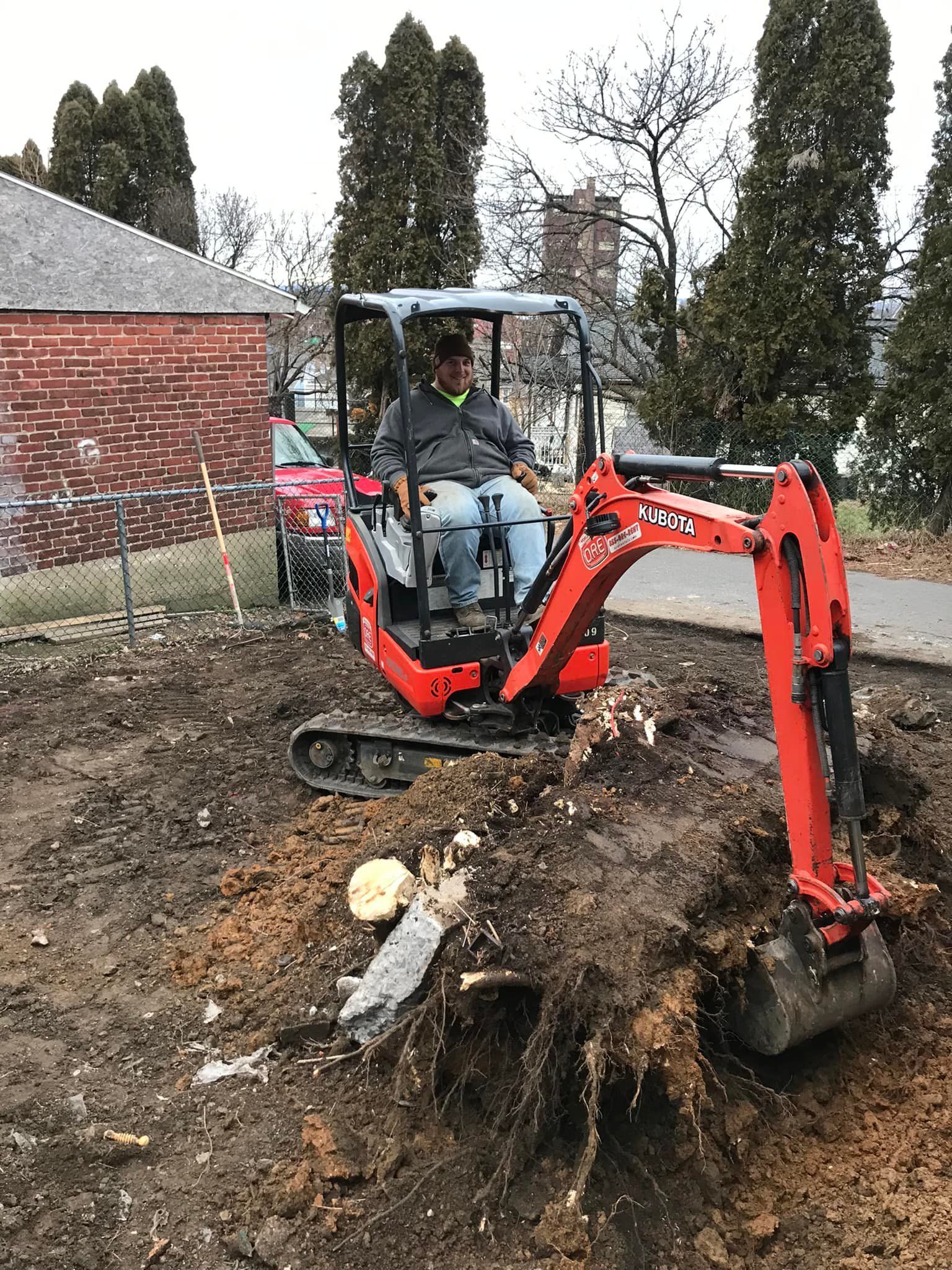 Man operating a small, orange excavator in a yard with exposed dirt and tree roots.