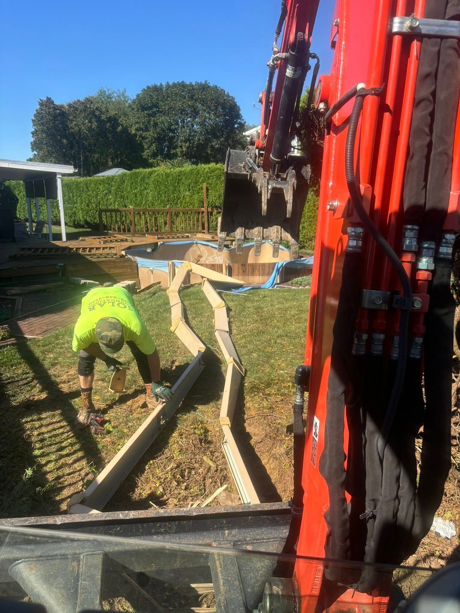 Worker in neon vest building wooden border near excavator in backyard.