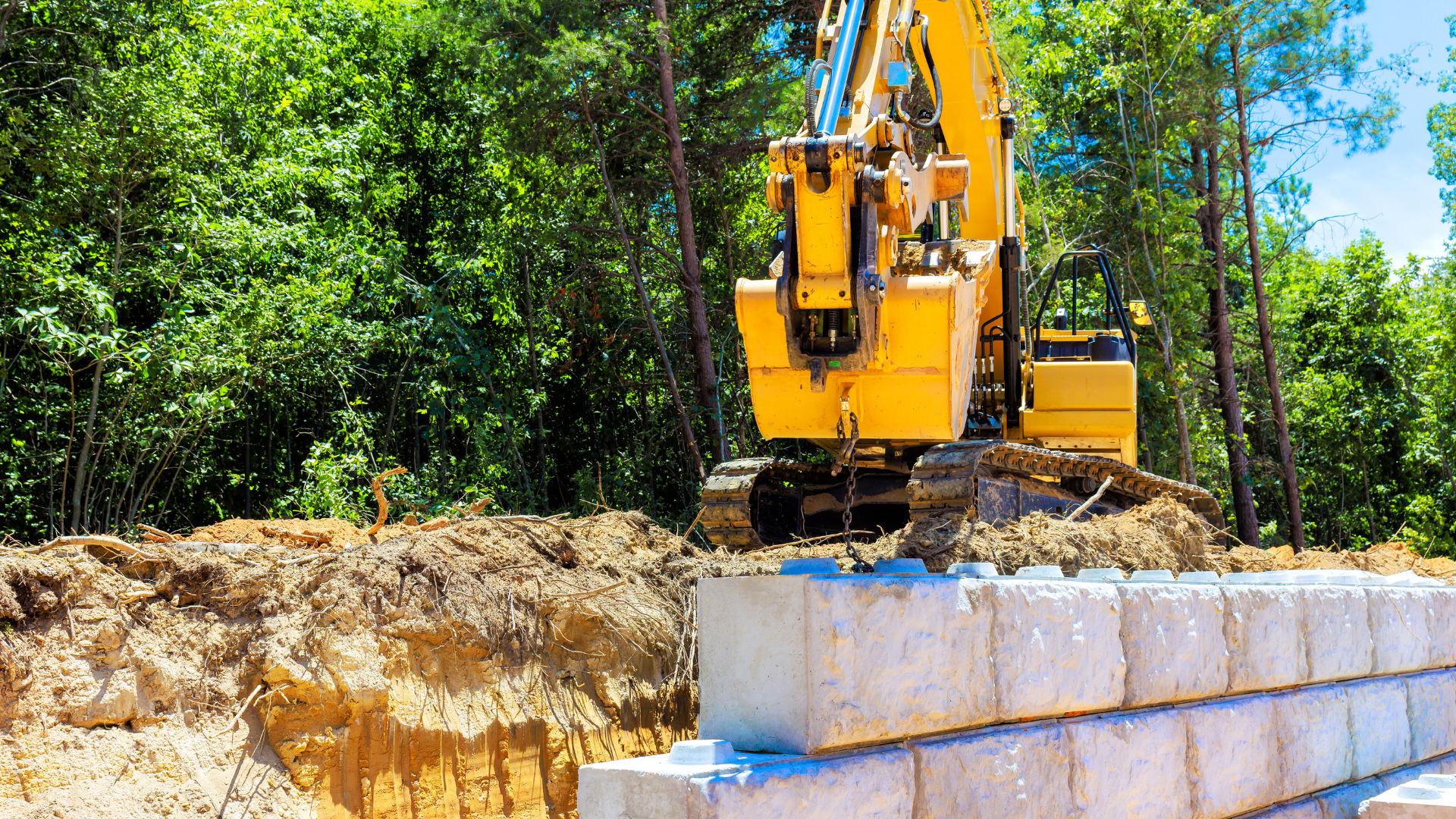 Yellow excavator building a retaining wall with concrete blocks near trees.
