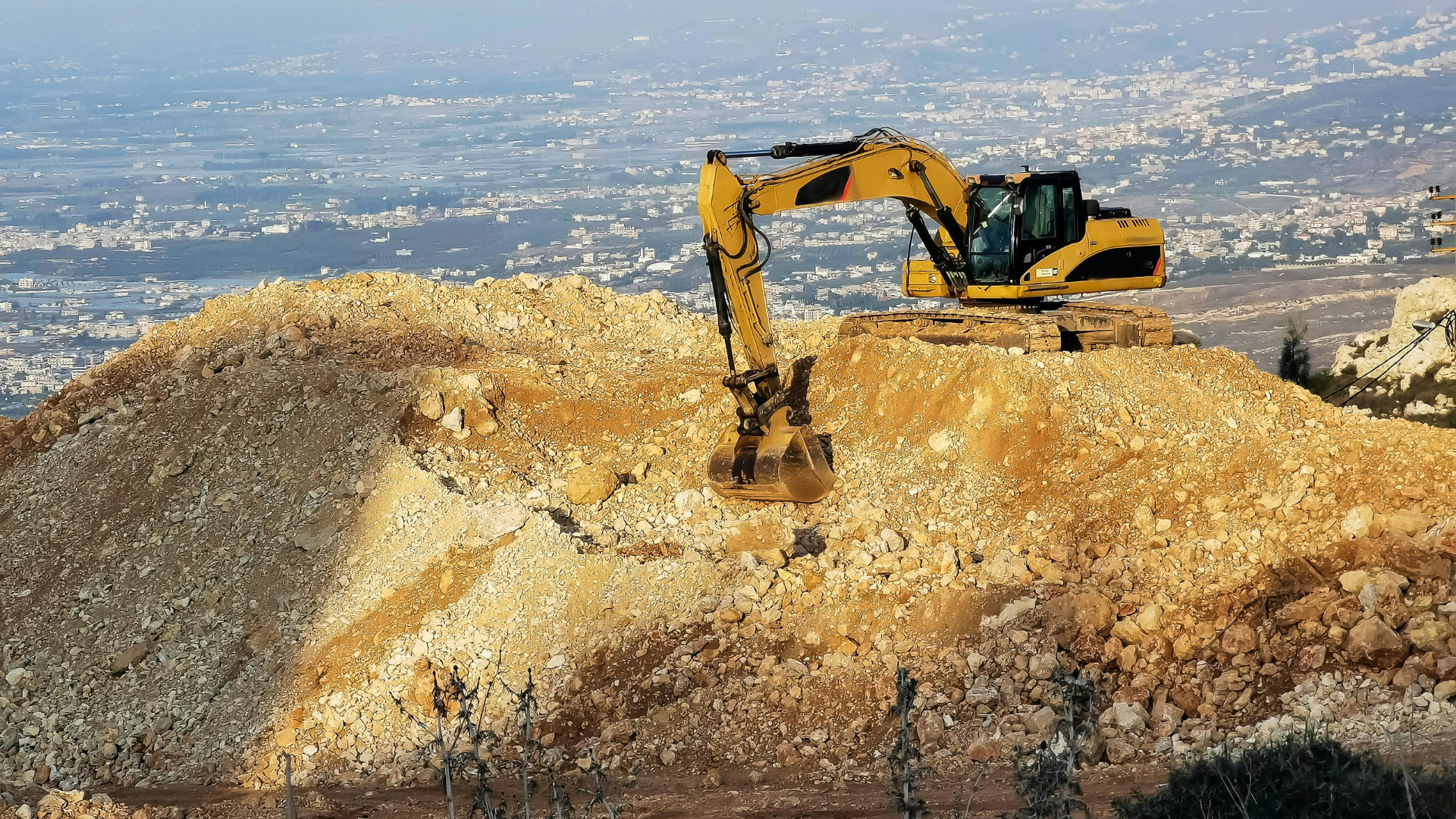 Excavator on a rocky hillside, removing earth. Distant cityscape in the background.