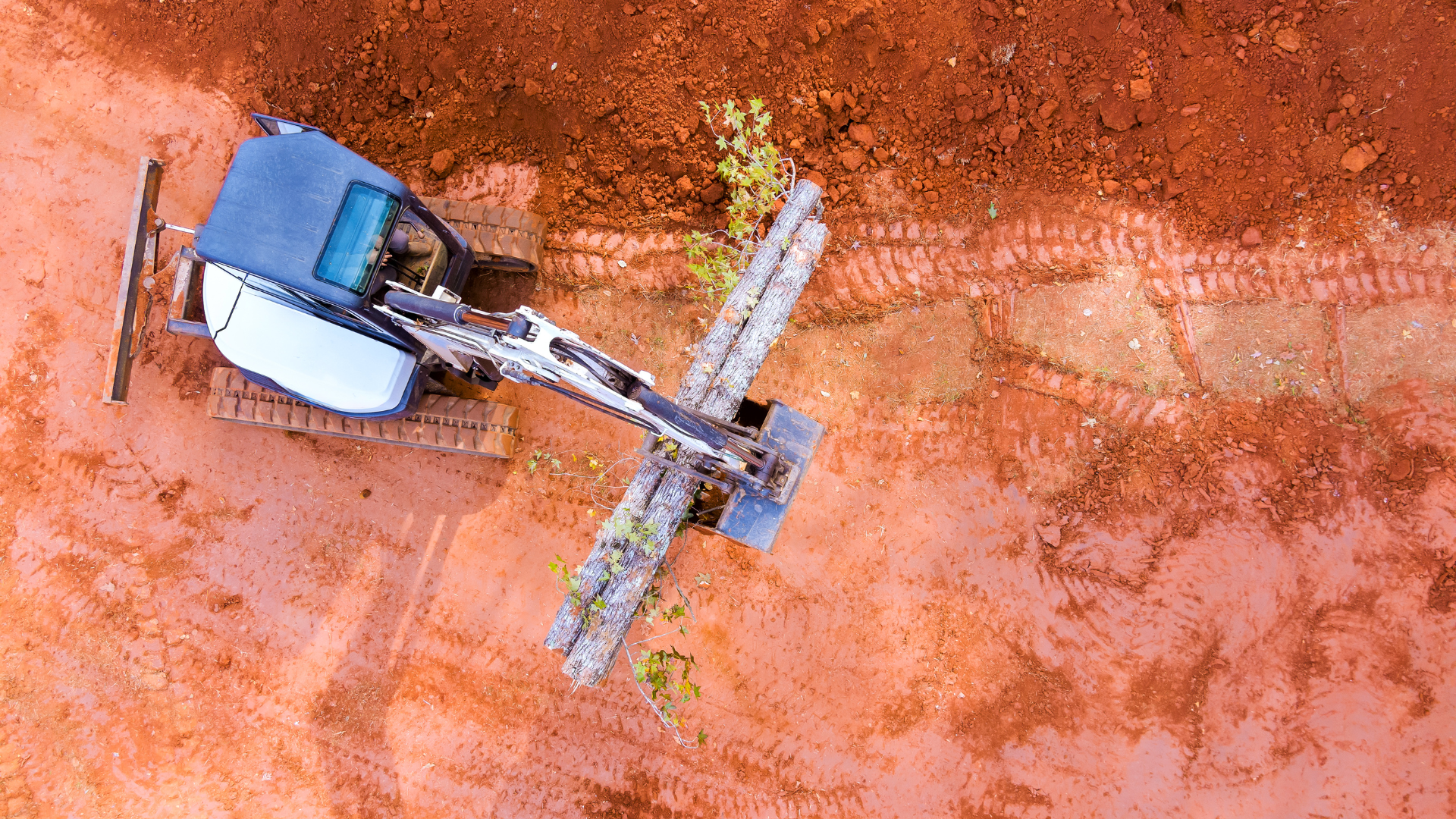 Overhead view: Excavator on red soil, some logs in front.