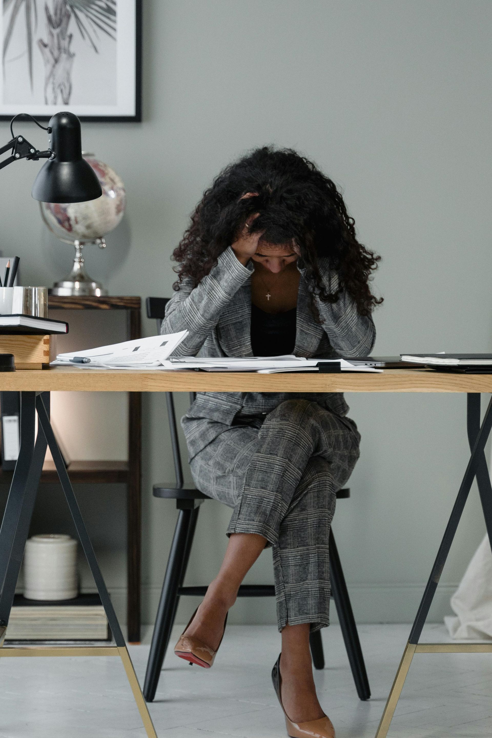 Woman in gray suit stressed at desk, head in hands.