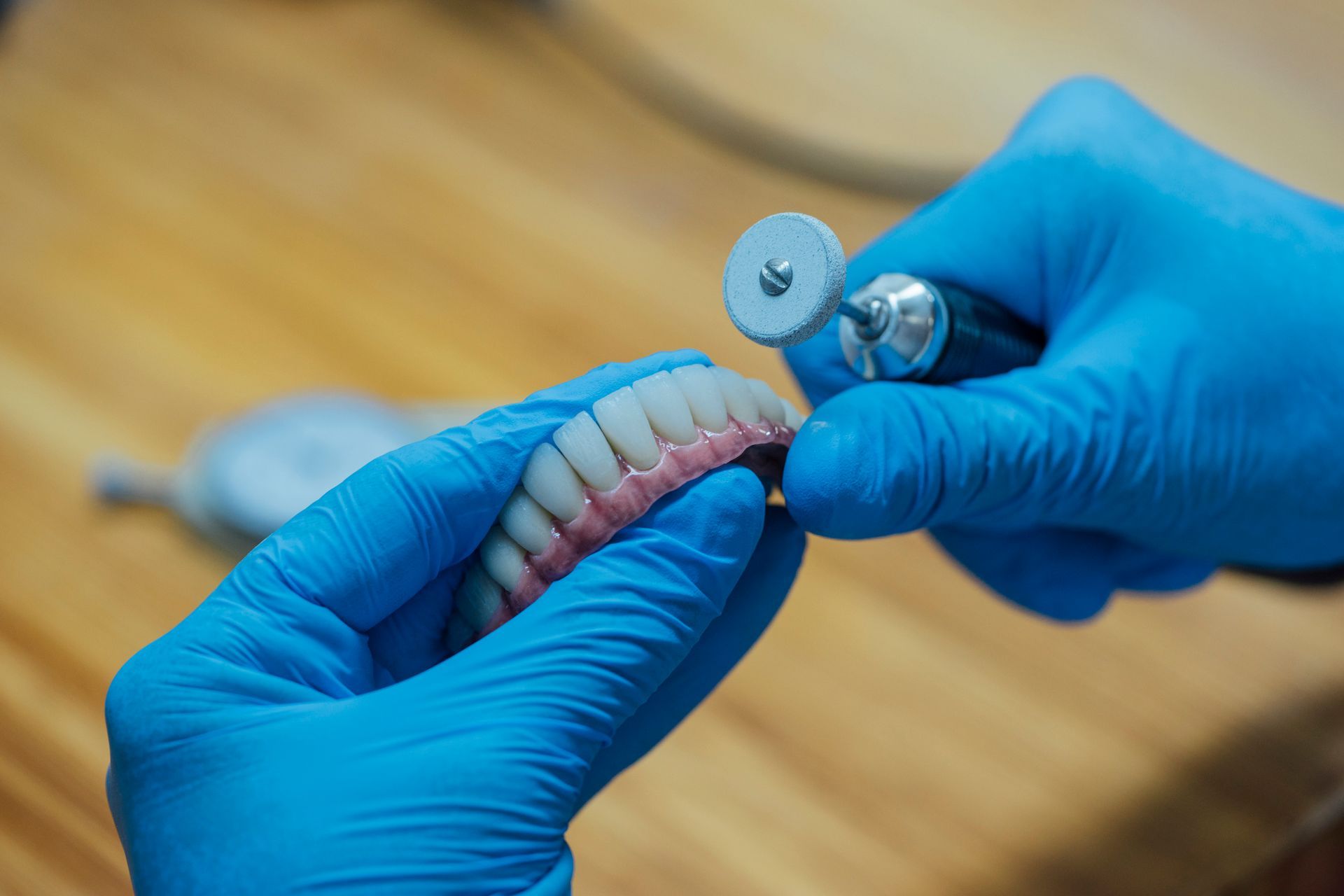 Hands in blue gloves polishing a set of false teeth with a small motorized tool.