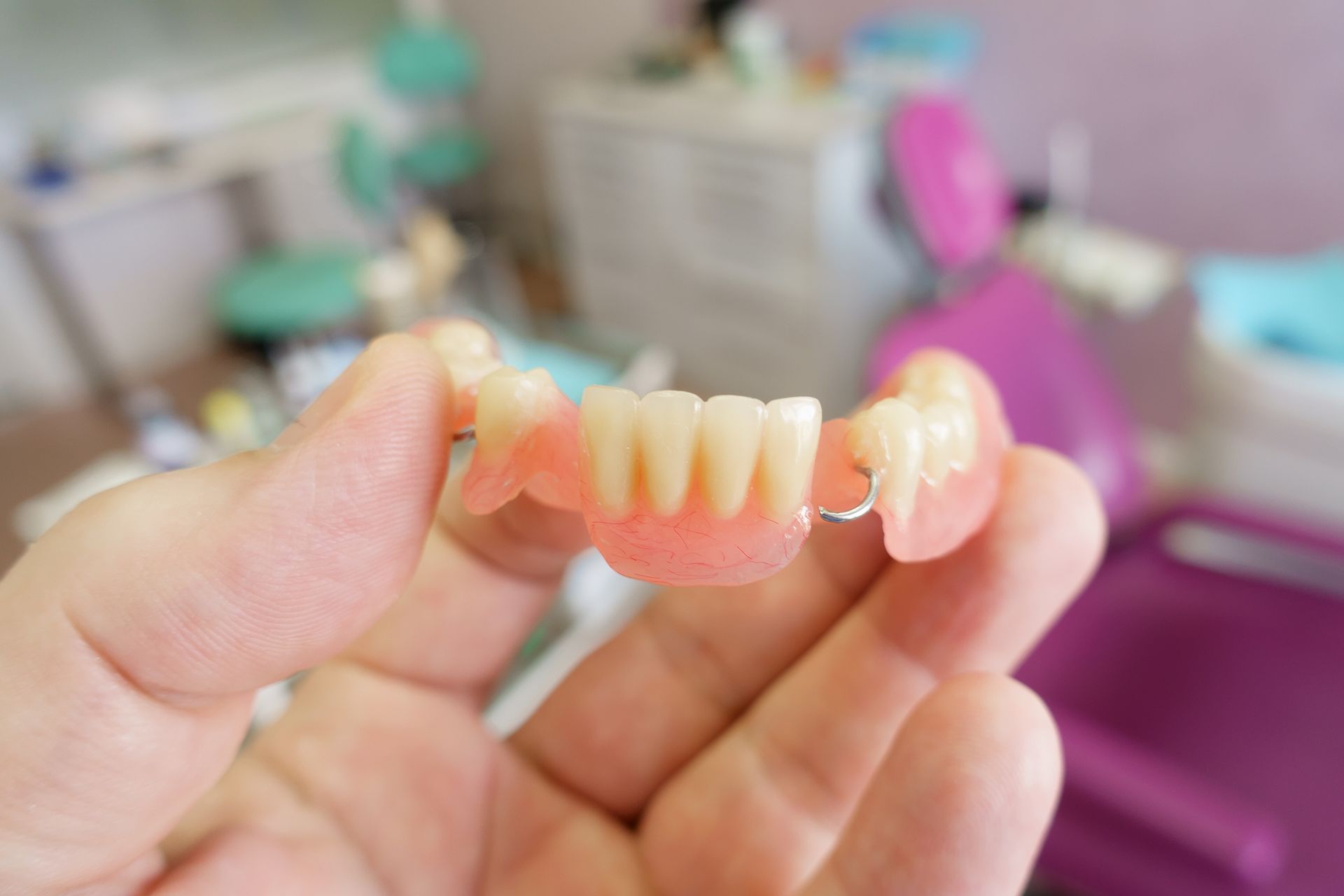 Hand holding a partial denture with pink gums and artificial teeth in a dental office.