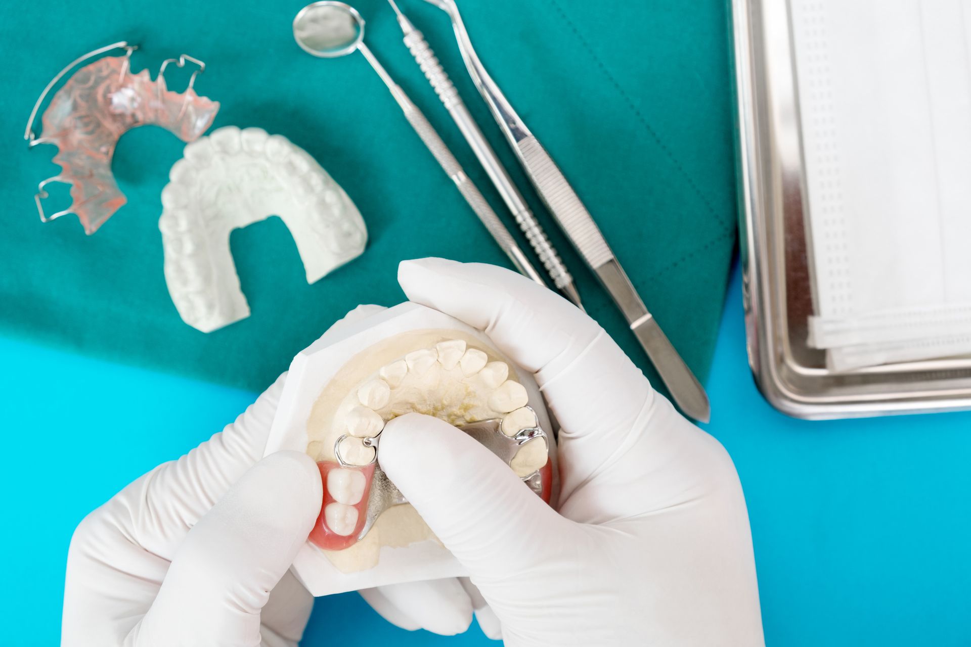 Hands wearing gloves holding a dental mold with dentures, surrounded by tools on a blue and green surface.