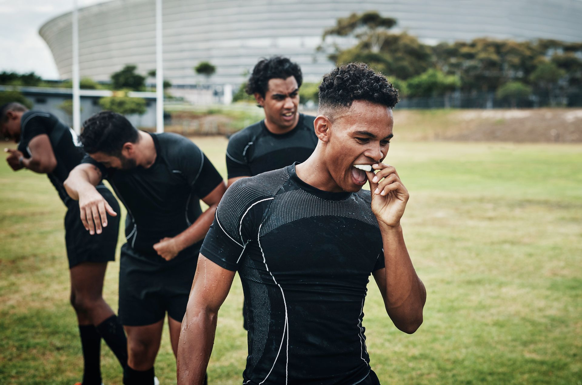 Rugby players in black jerseys celebrate on a green field. One bites a mouthguard, smiling.