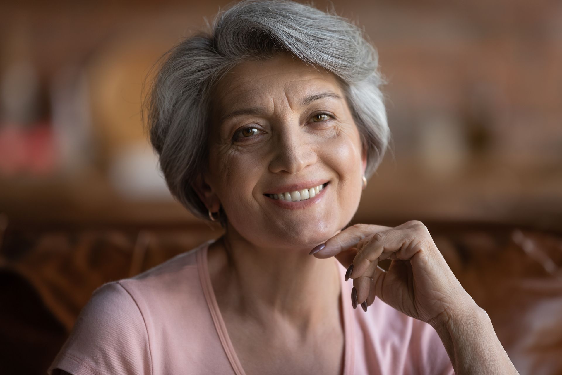 Smiling older woman with gray hair, wearing a pink shirt, resting chin on her hand.