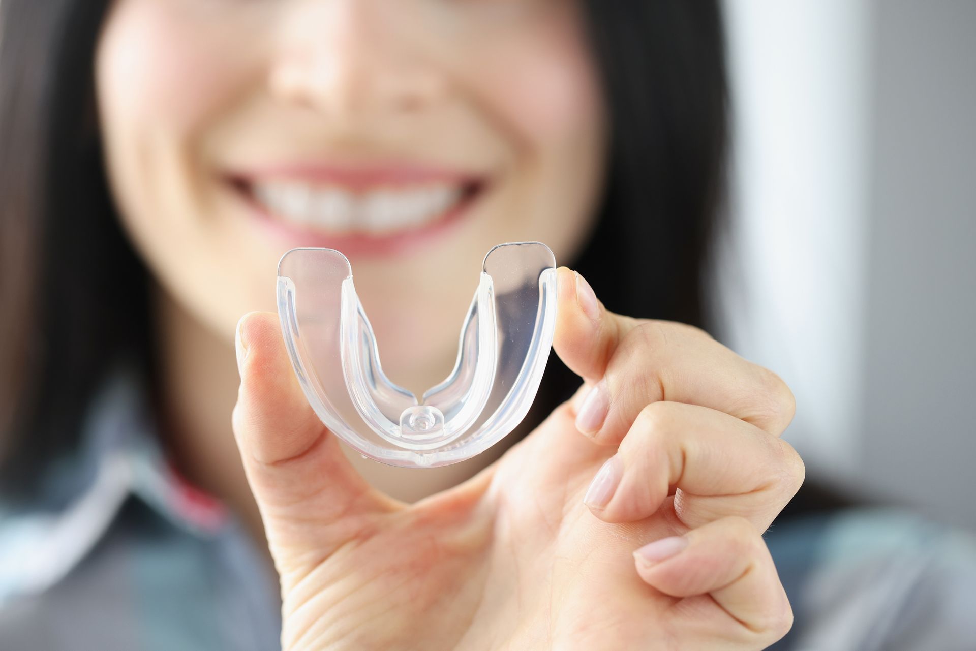 Woman holding a clear, U-shaped mouthguard, smiling in a well-lit room.