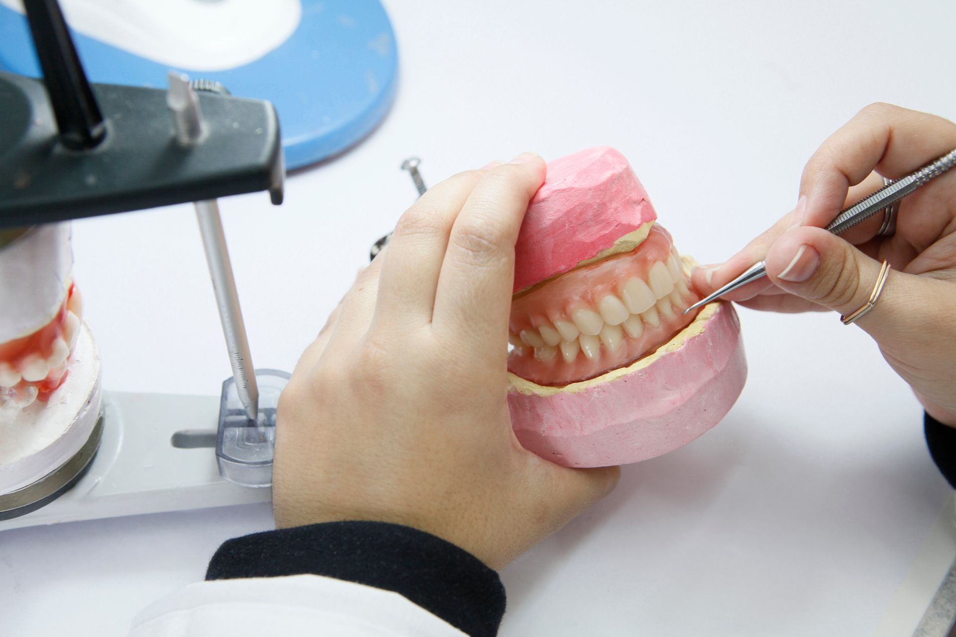 Hands of a dental technician working on a pink model of teeth with a metal tool.
