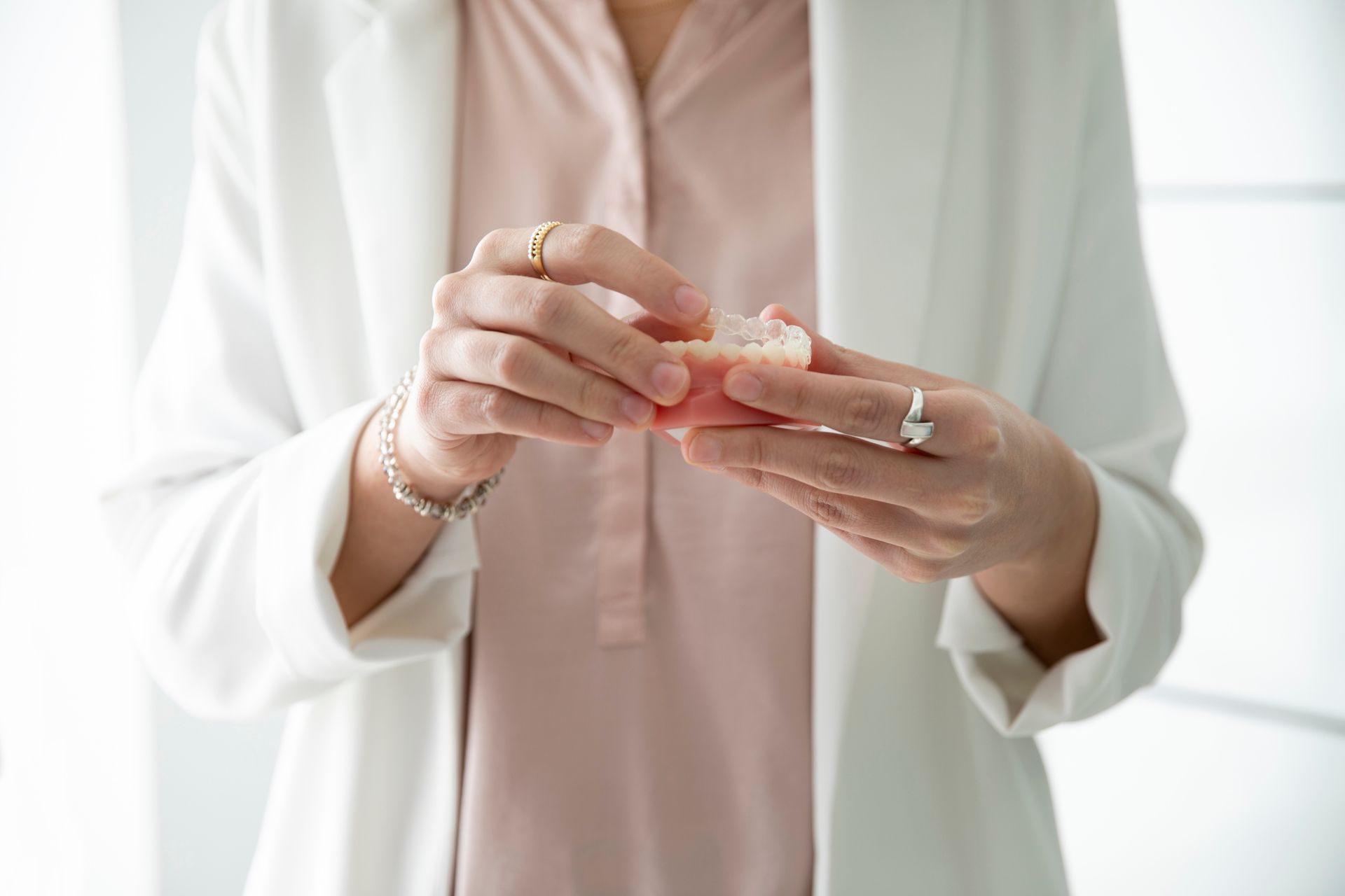 Woman in white blazer and pink shirt holding a set of dentures, wearing rings.
