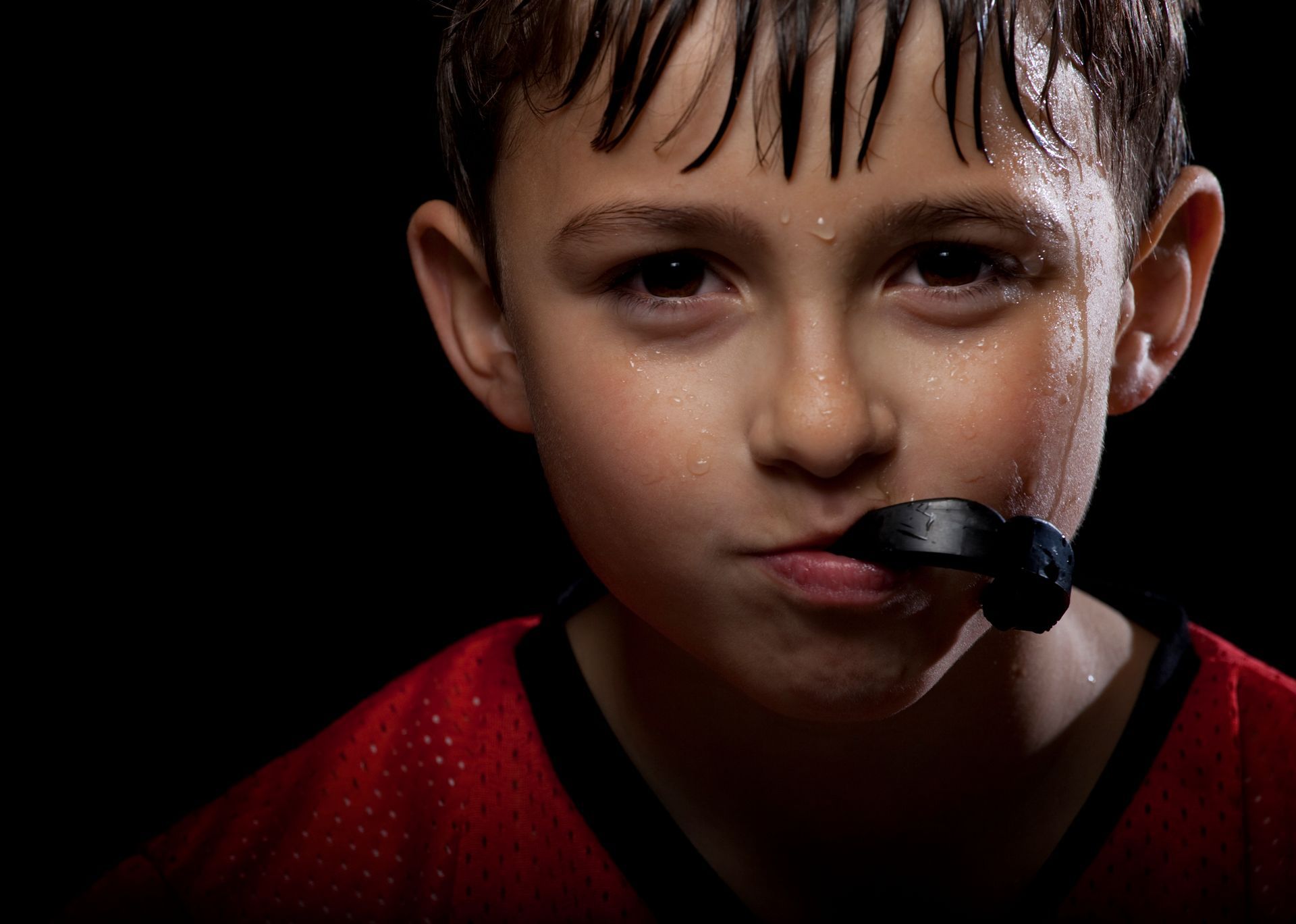 Boy in red jersey with a mouthguard, sweating with intense gaze against a black background.