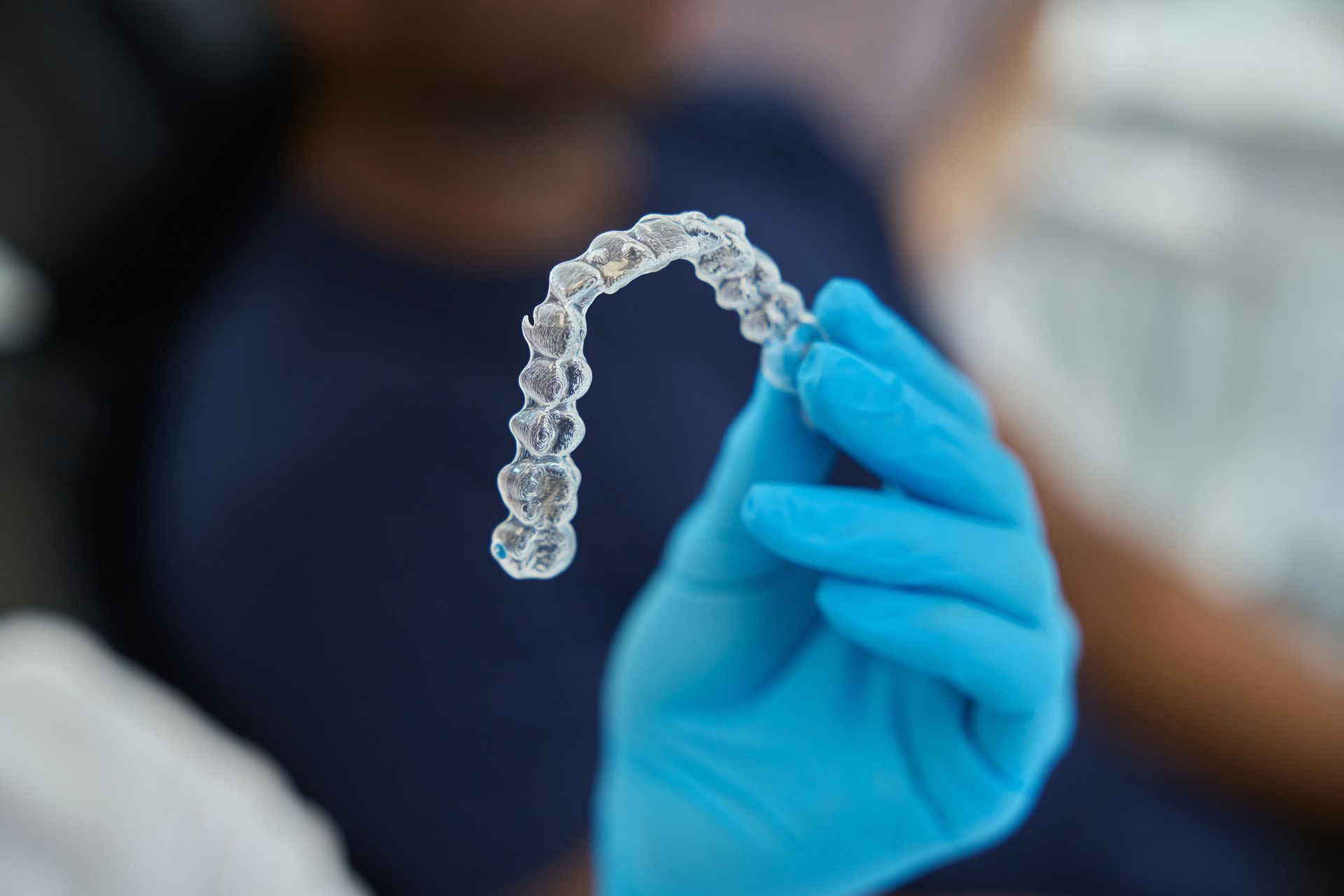 Gloved hand holds a clear Invisalign aligner, in a dental office setting.