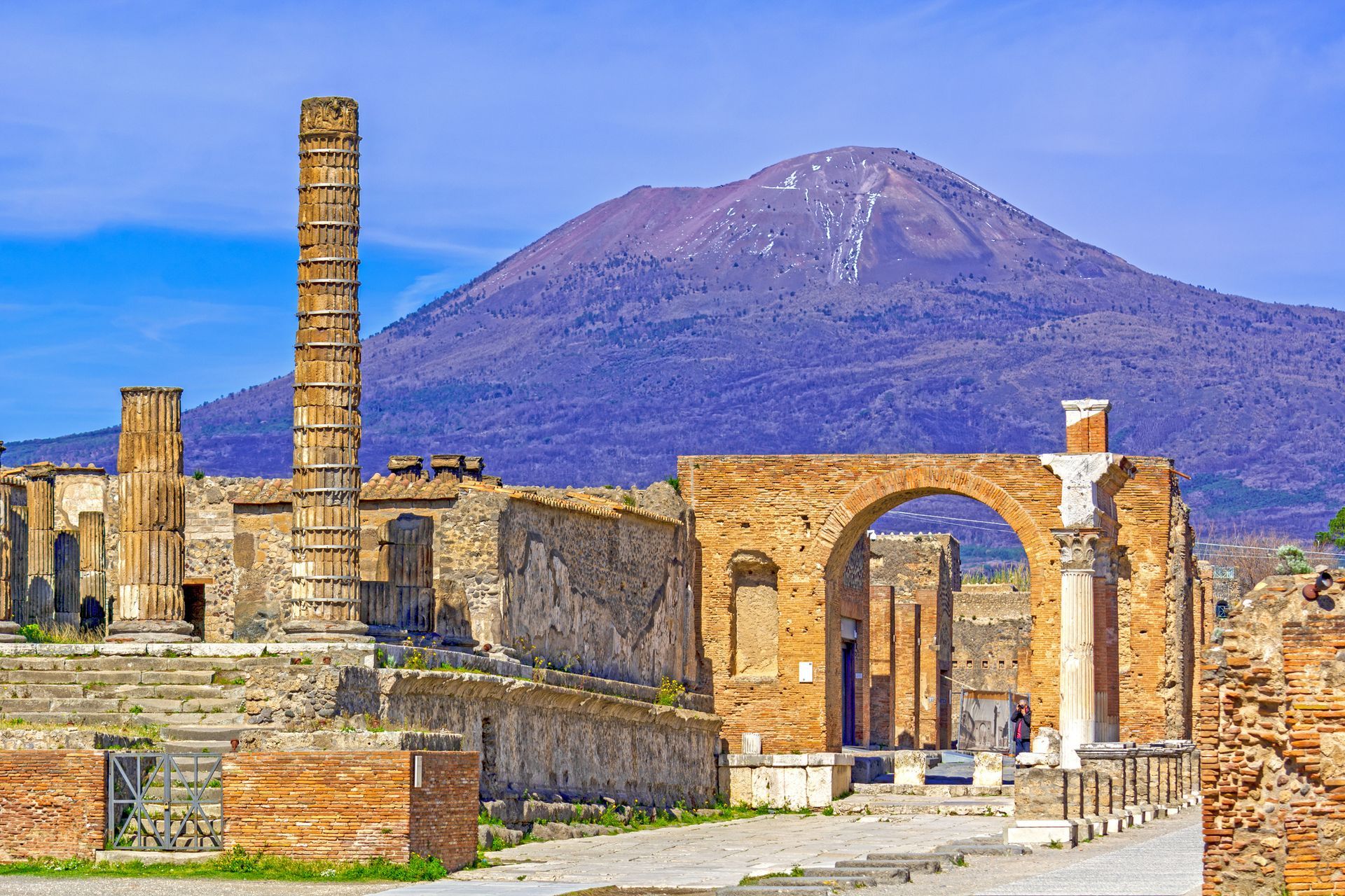 Rovine di Pompei con il Vesuvio sullo sfondo. Le strutture dell'antica città sono in vari stati di degrado e presentano una tonalità arancione-marrone.