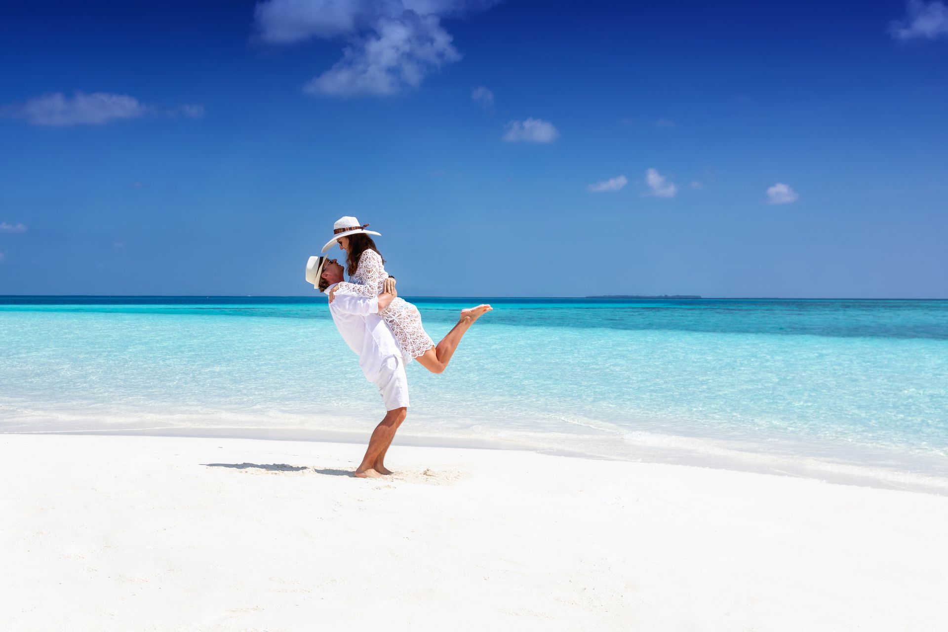 Un uomo solleva una donna su una spiaggia di sabbia bianca, entrambi vestiti di bianco. Acqua turchese e cielo azzurro sullo sfondo.
