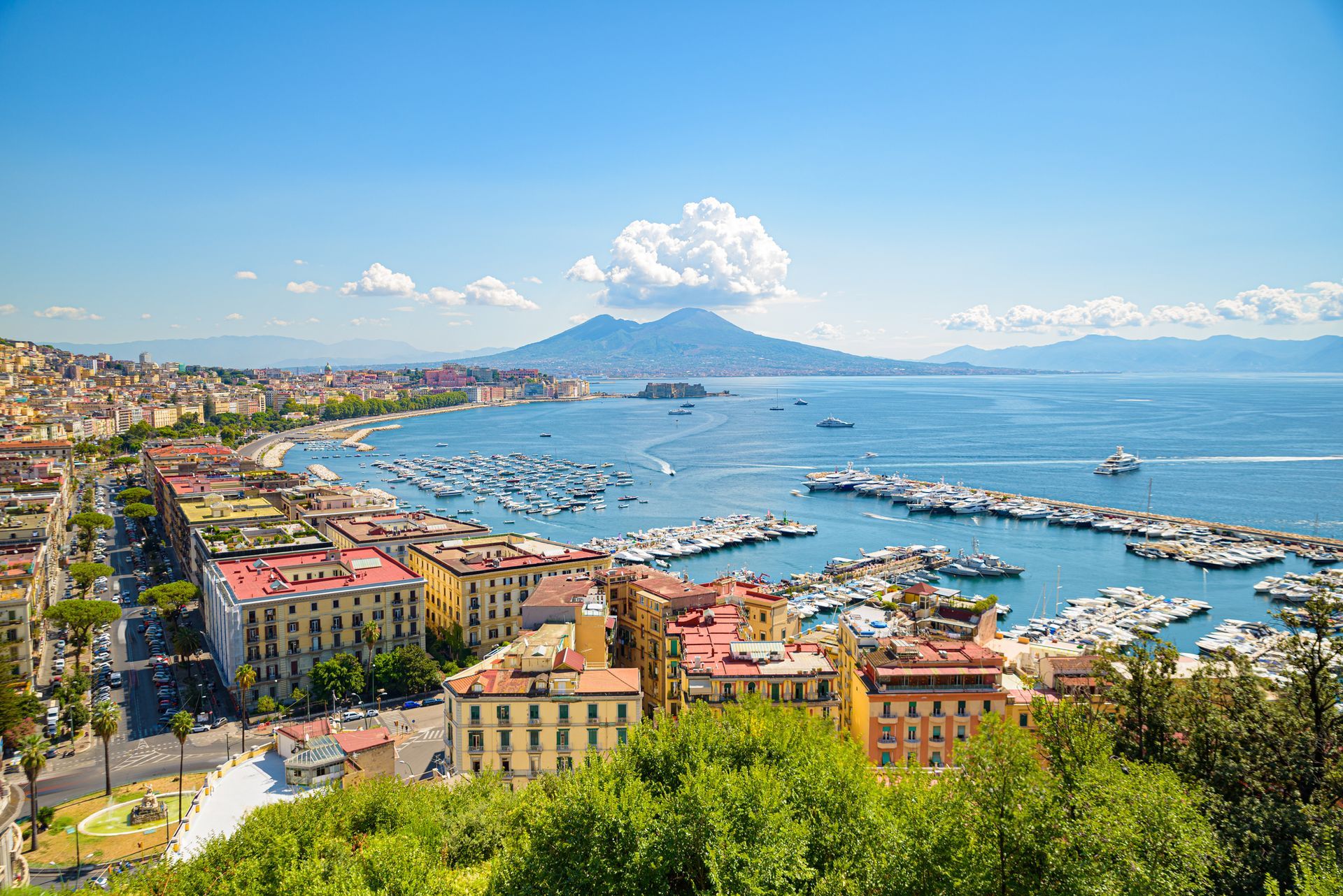 Vista costiera di Napoli, Italia, con edifici lungo la riva, barche nel porto e il Vesuvio sullo sfondo sotto un cielo azzurro.