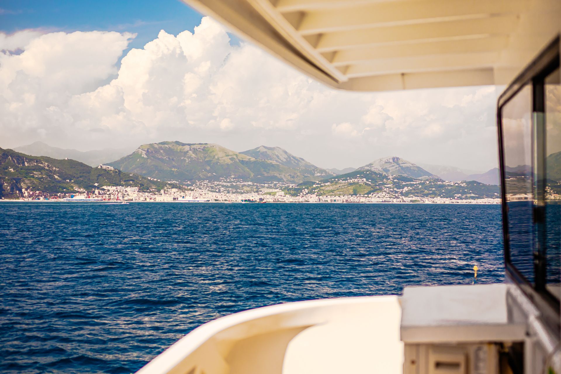 Vista da una barca sulle acque blu verso una città costiera con montagne sotto un cielo parzialmente nuvoloso.