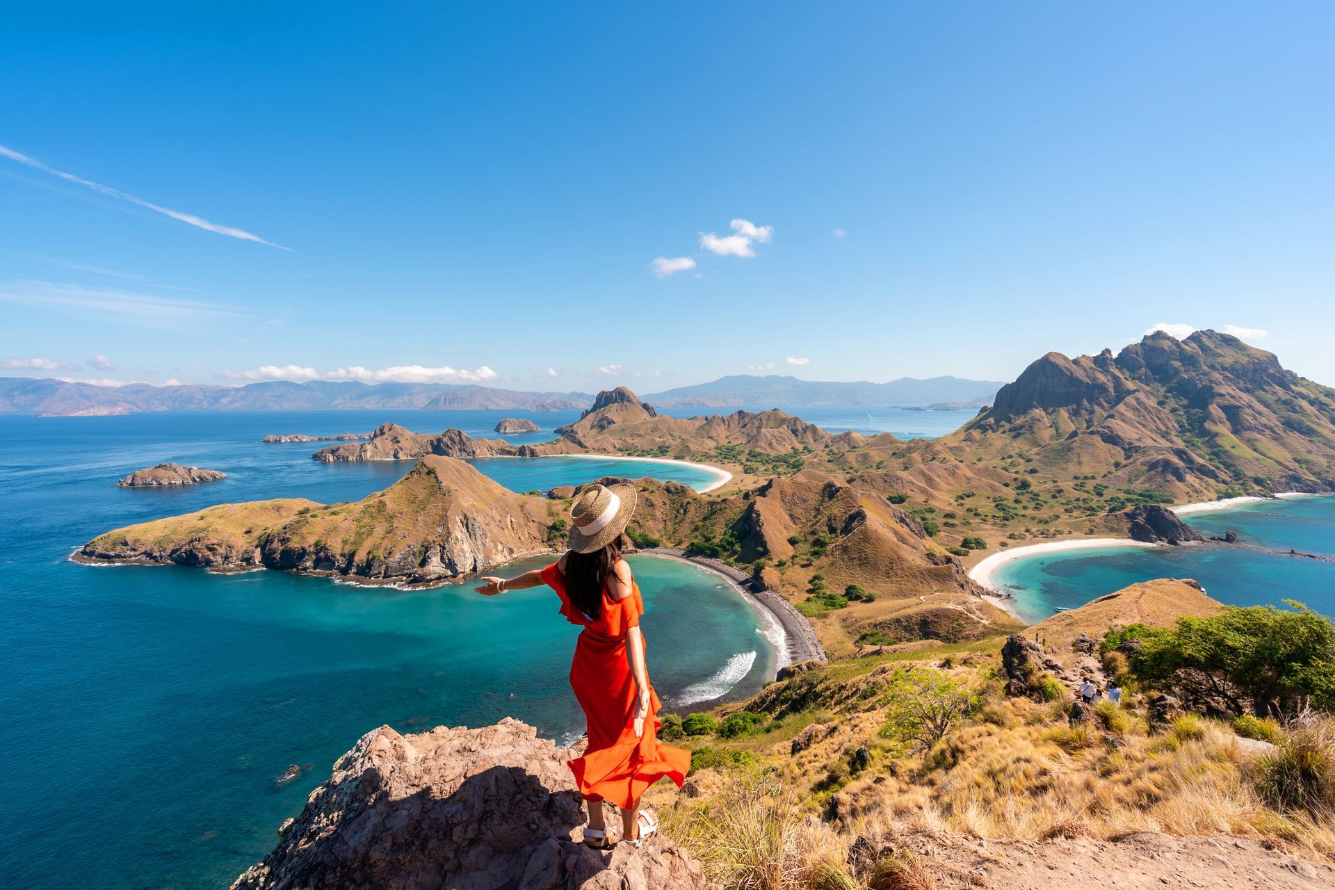 Donna con un abito rosso e le braccia tese, in piedi su una scogliera che domina l'oceano costellato di isole. Acqua e cielo di un azzurro intenso.