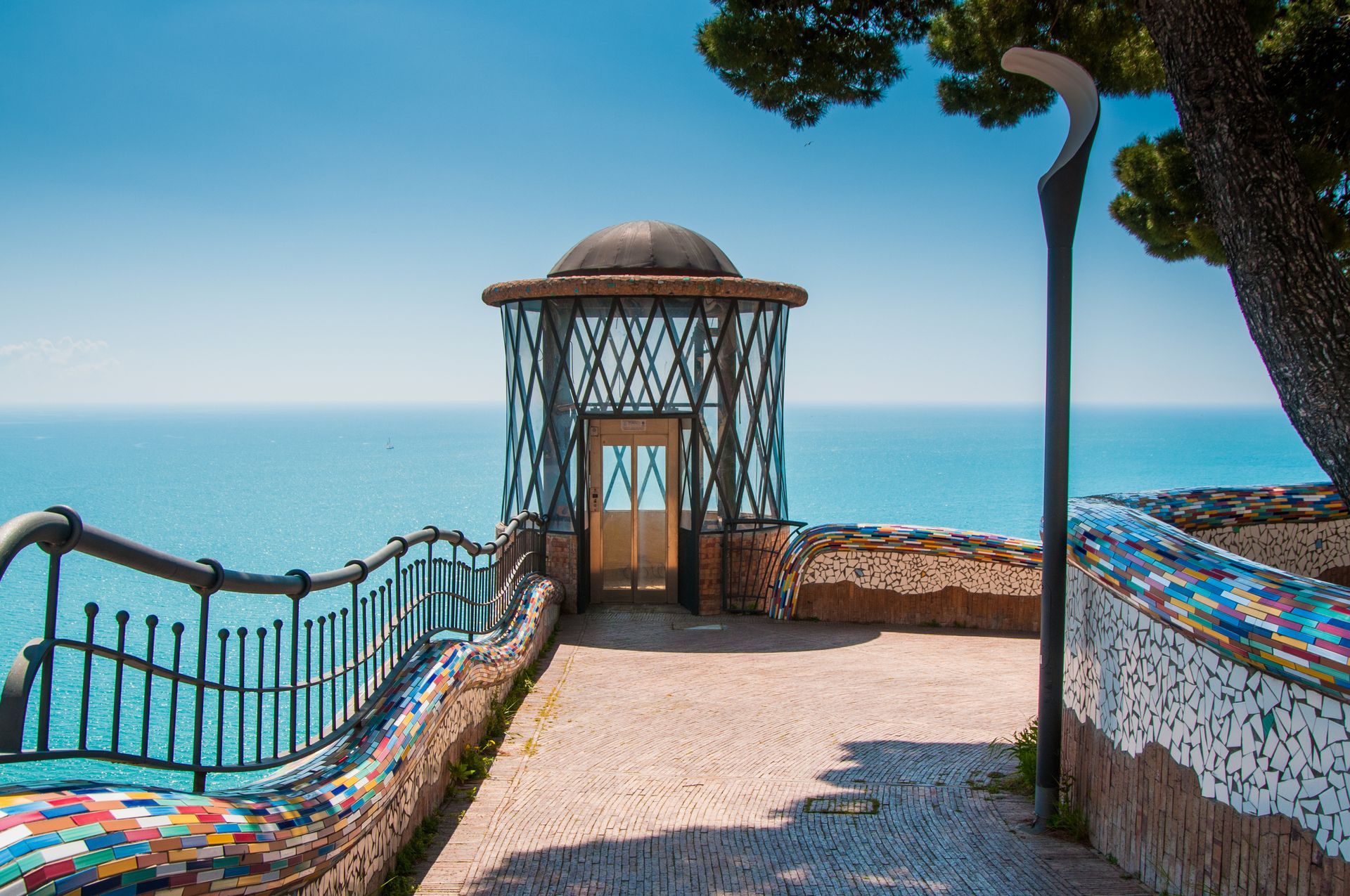 Gazebo con vista sull'oceano, con un sentiero rivestito di mosaici e un albero sulla destra, sotto un cielo azzurro e terso.
