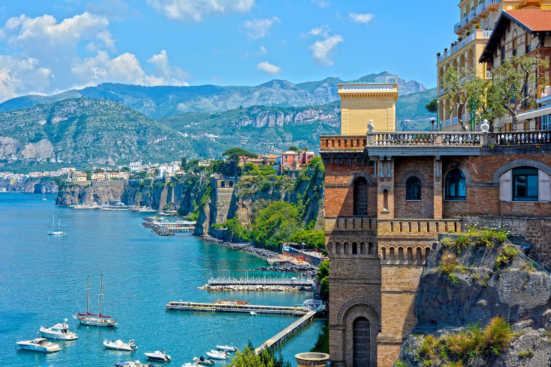 Vista costiera di Sorrento, Italia. Edifici arroccati sulle scogliere a picco sul mare blu, con barche e montagne sullo sfondo.