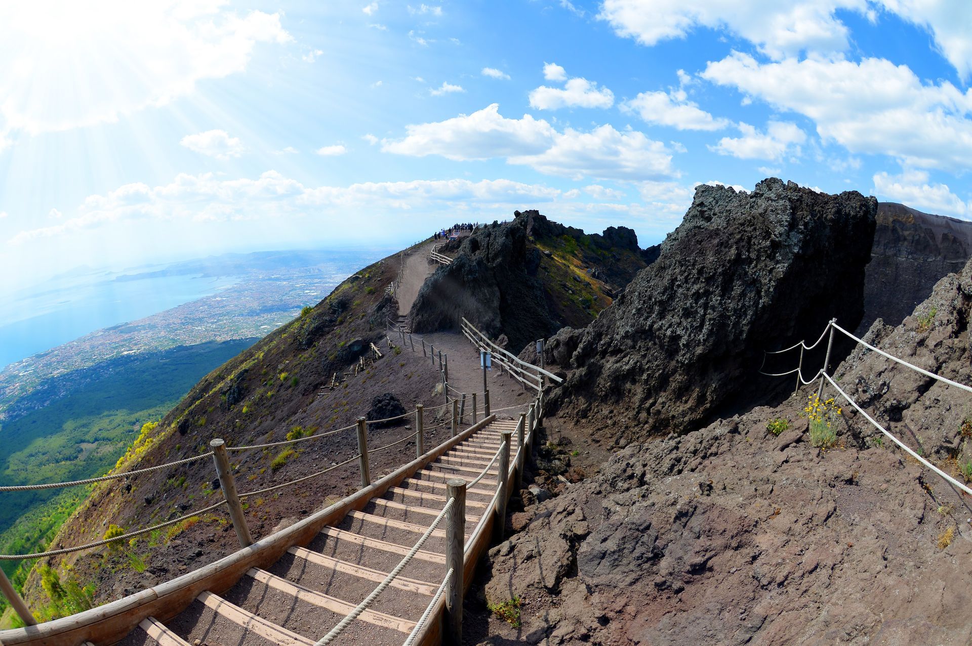 Una scalinata conduce sul terreno aspro e roccioso del Vesuvio, che domina una città costiera sotto un cielo azzurro.