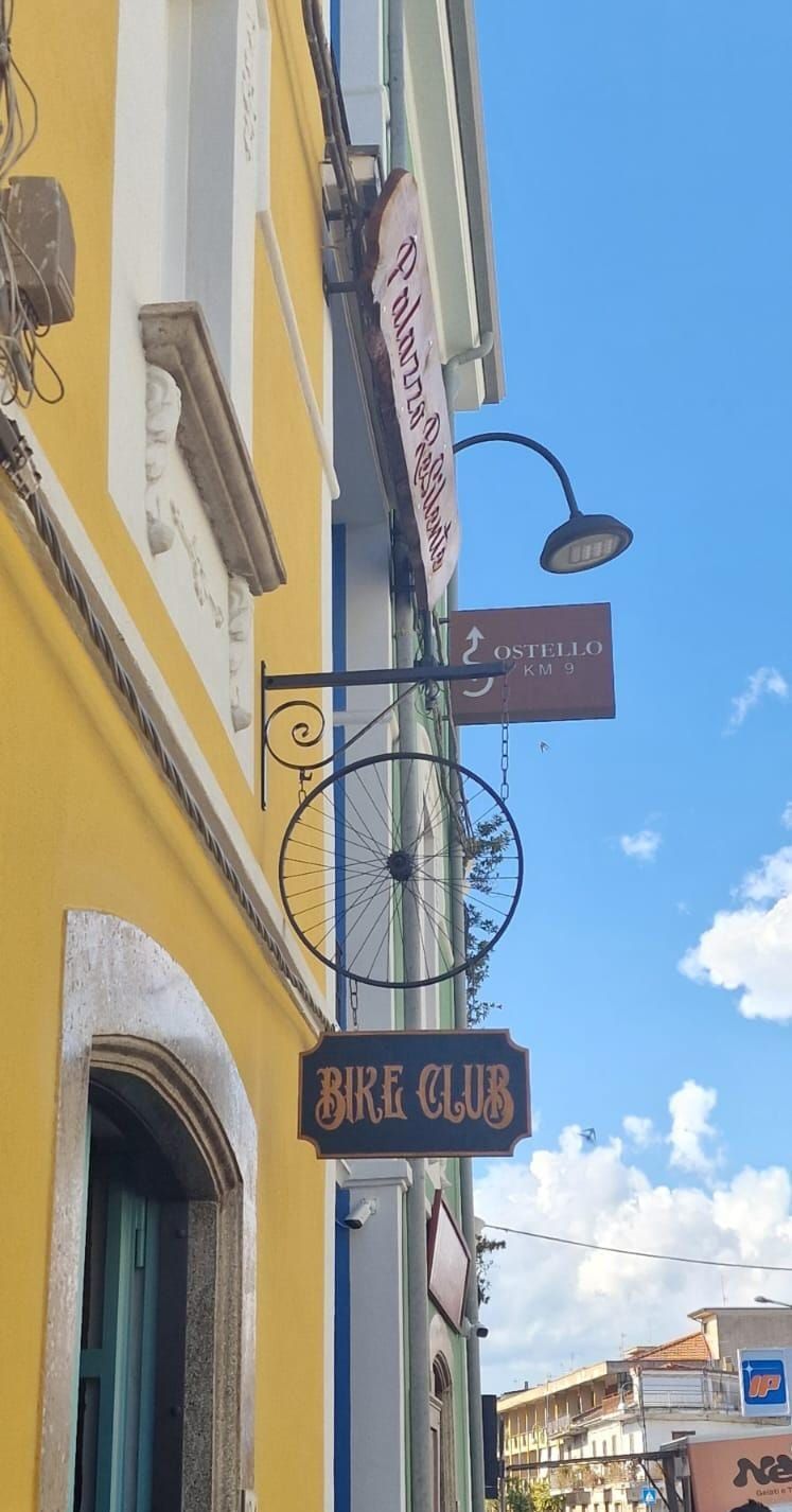 Edificio giallo con insegne di un club e di un ristorante; dettagli in ferro battuto nero con il disegno di una ruota di bicicletta.