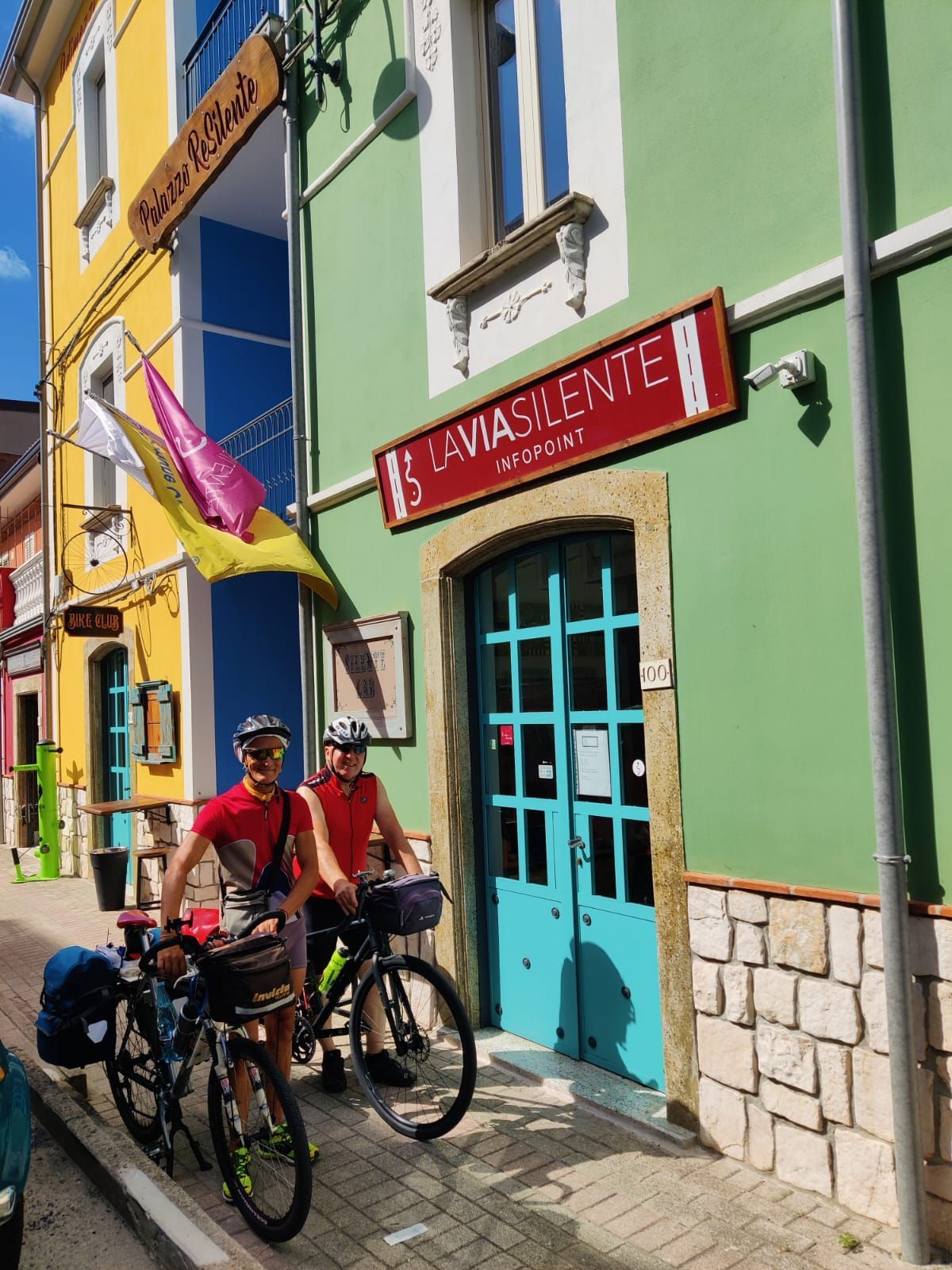 Due ciclisti e biciclette davanti a un edificio verde con un cartello rosso che dice