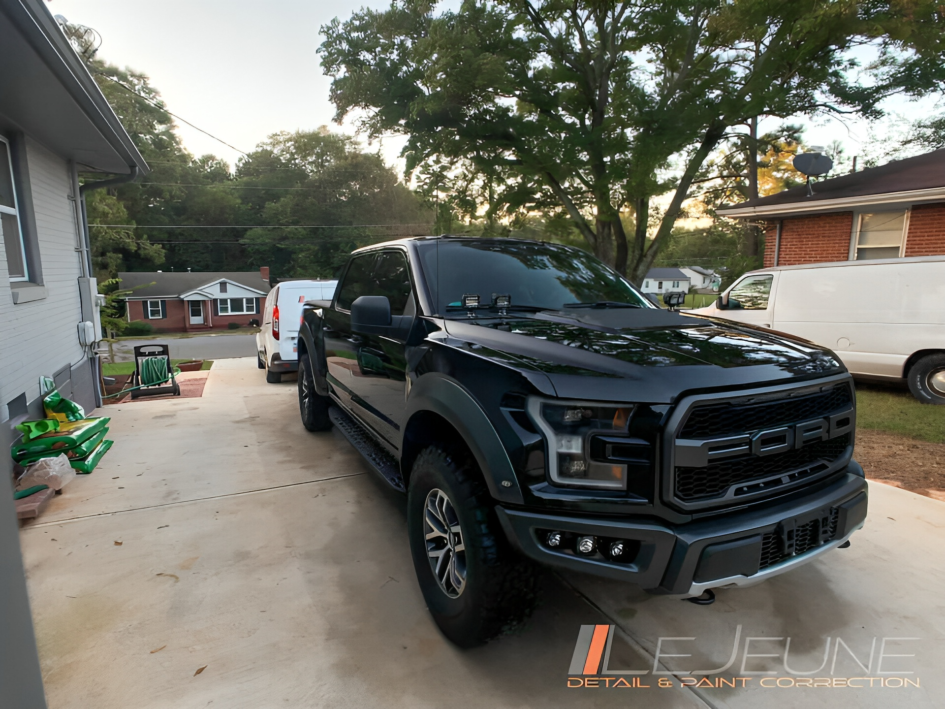 Black Ford Raptor truck parked in a driveway with a white van and house in the background.