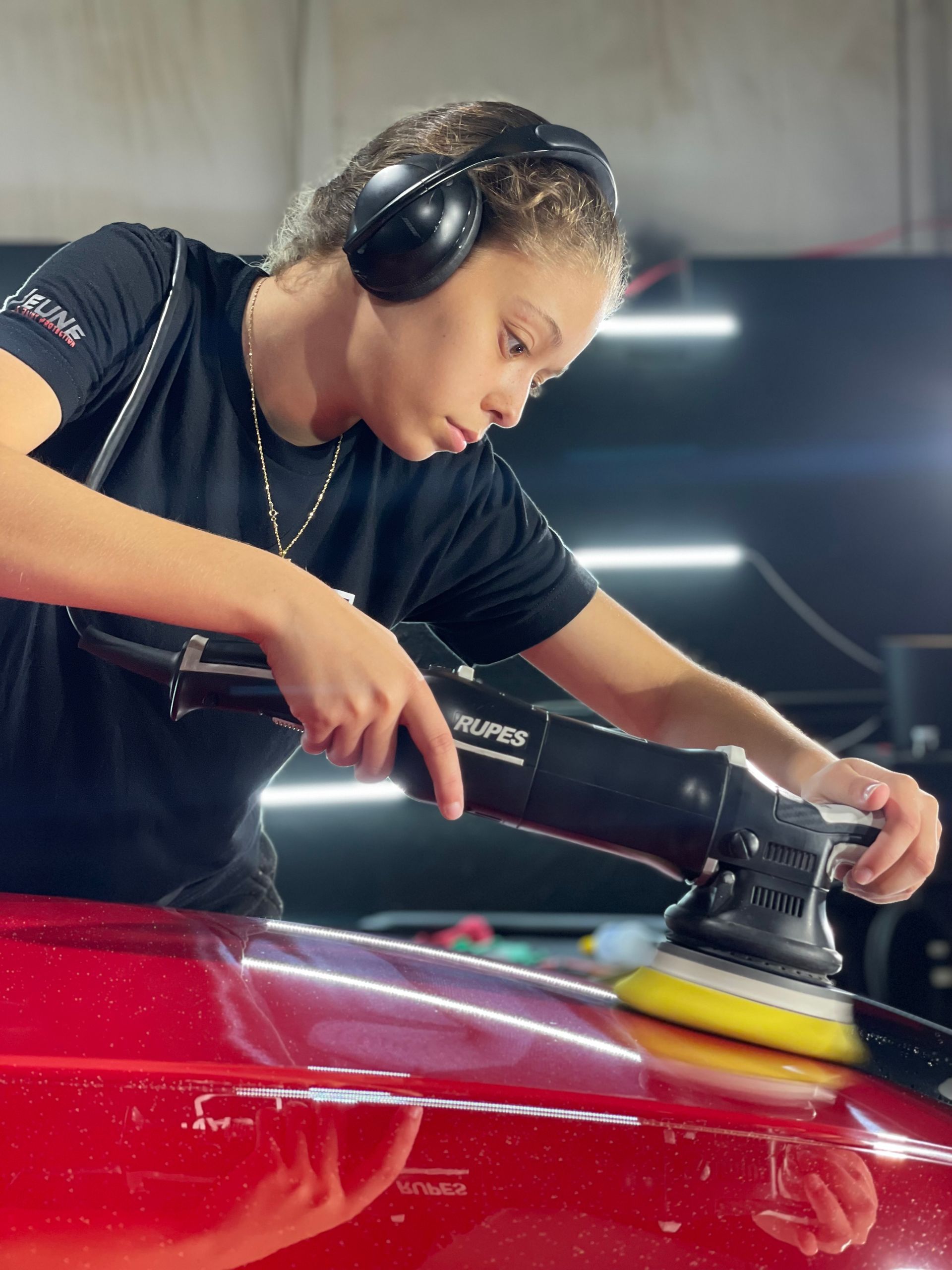 A woman wearing headphones is polishing a red car with a machine.