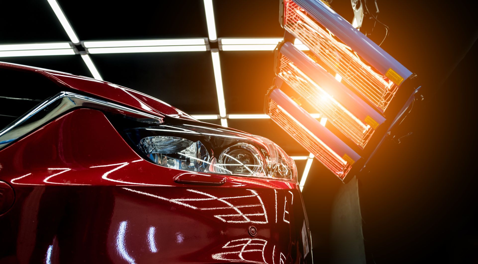 Red car under infrared paint drying lamp in a brightly lit body shop.