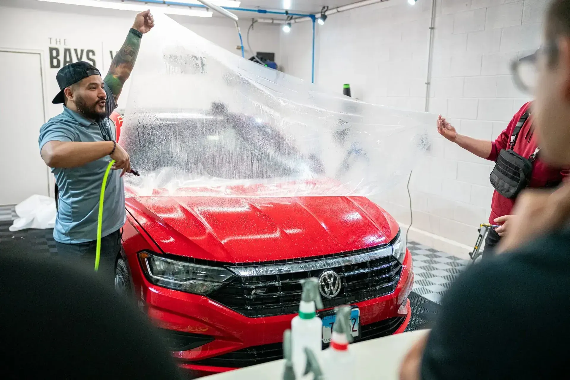 A man is covering a red car with a plastic sheet.