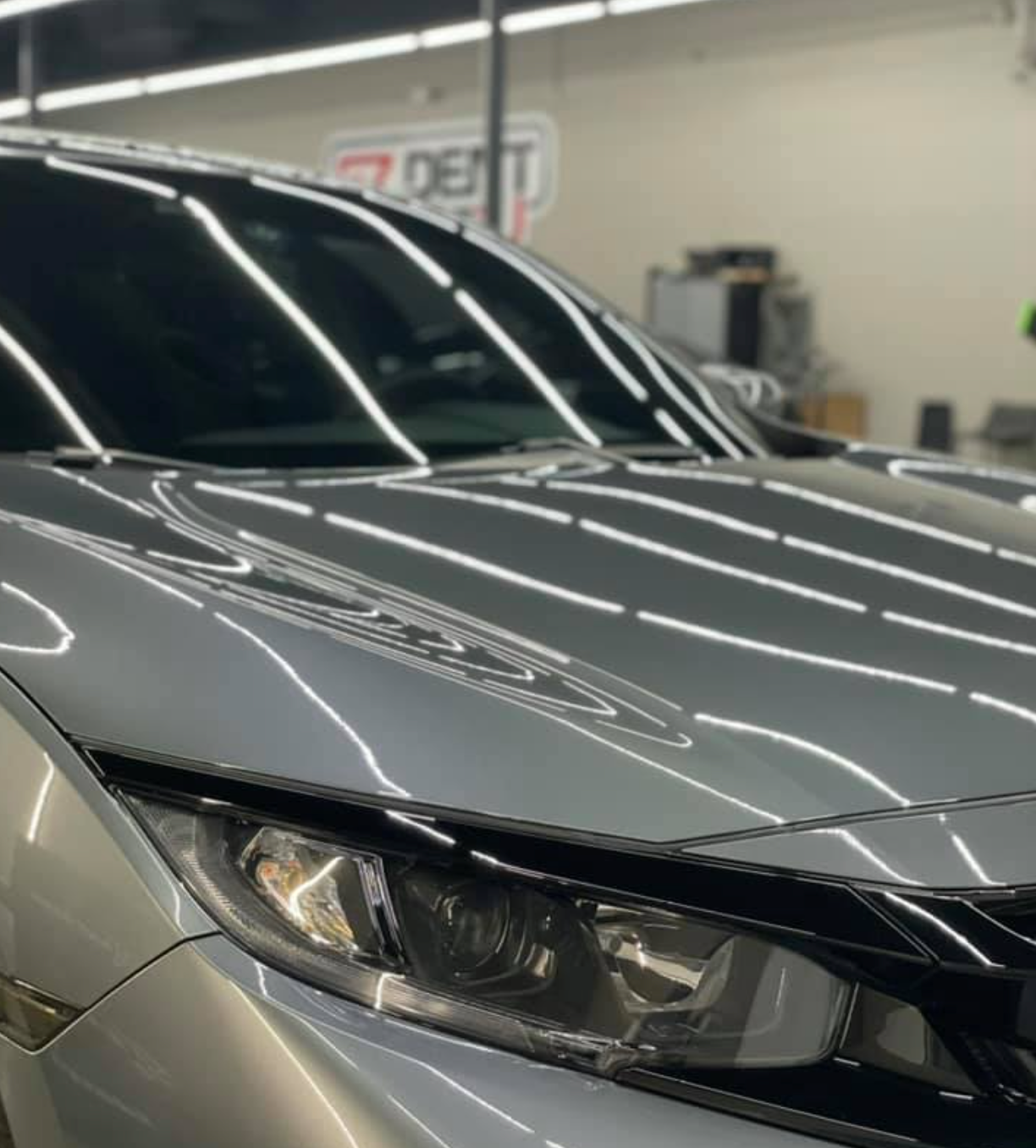 Silver car's shiny hood and headlights with a dark tinted windshield reflecting lights in a shop.