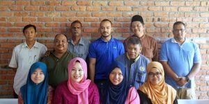 Group of diverse people posing in front of a brick wall. Some are wearing hijabs, others are smiling.