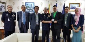 A group of seven people stand together in an office. Several men are wearing suits, and one man is holding a small card. A woman in a hijab is also in the group.