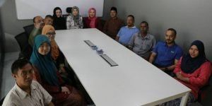 A group of people, some wearing hijabs, seated around a large white table in a meeting room.