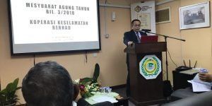 A man in a suit gives a presentation at a podium. A screen displays text about a cooperative.
