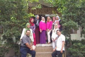A group of people, mostly women wearing hijabs and colorful clothing, pose on steps in front of a building with greenery.